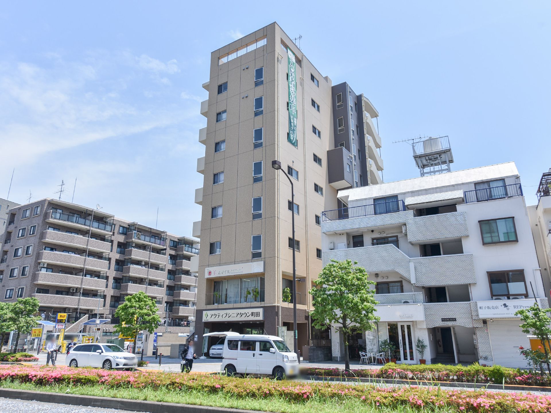 中央には高層階建ての建物があり、道路に面している。左側にはマンションがあり、その隣にはコインパーキングがある。