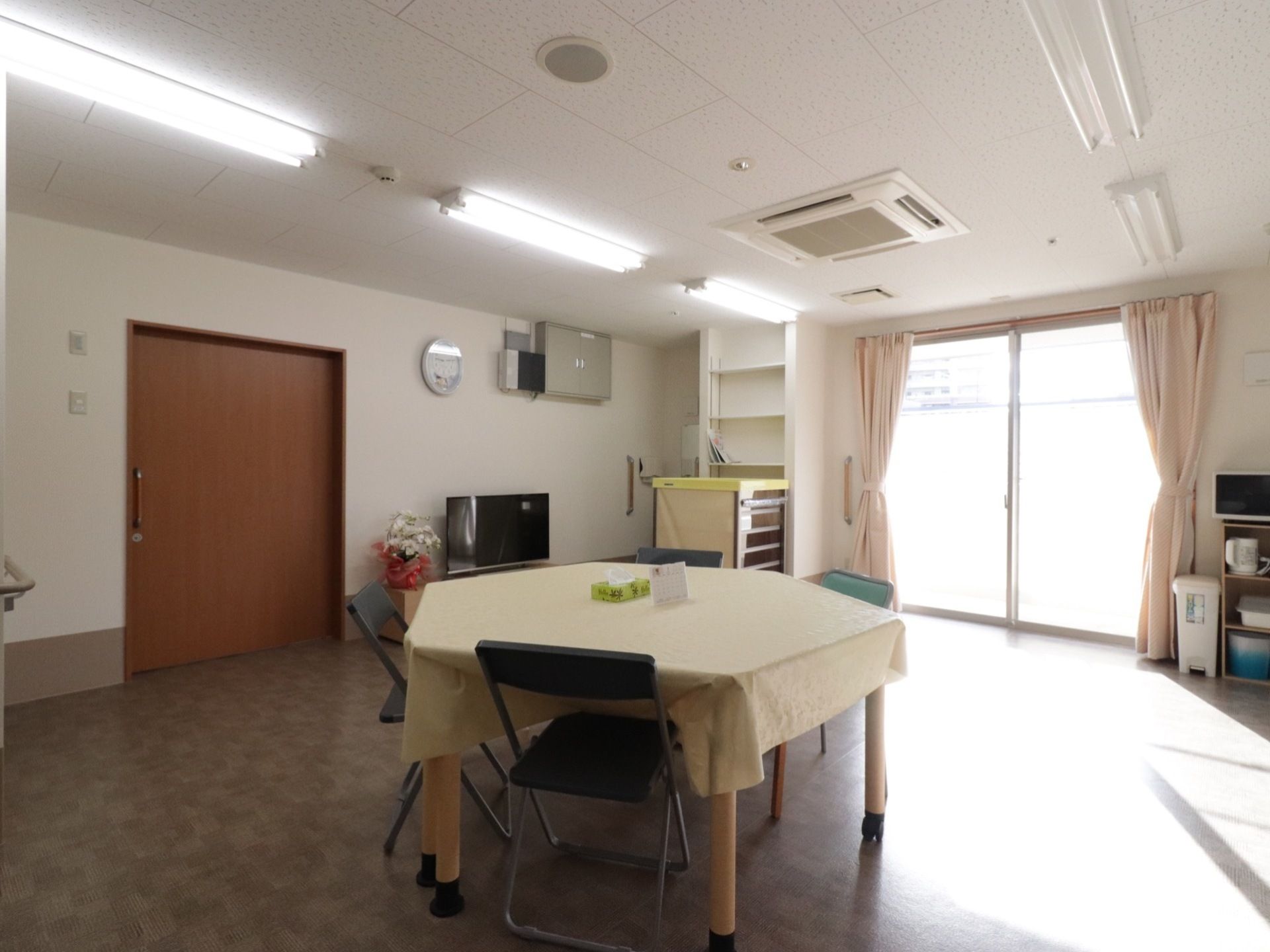 広々としたリビング。黄色いテーブルクロスのかかった4人掛けのテーブル。大きなガラス張りの窓からは外光が差し込んでいる。