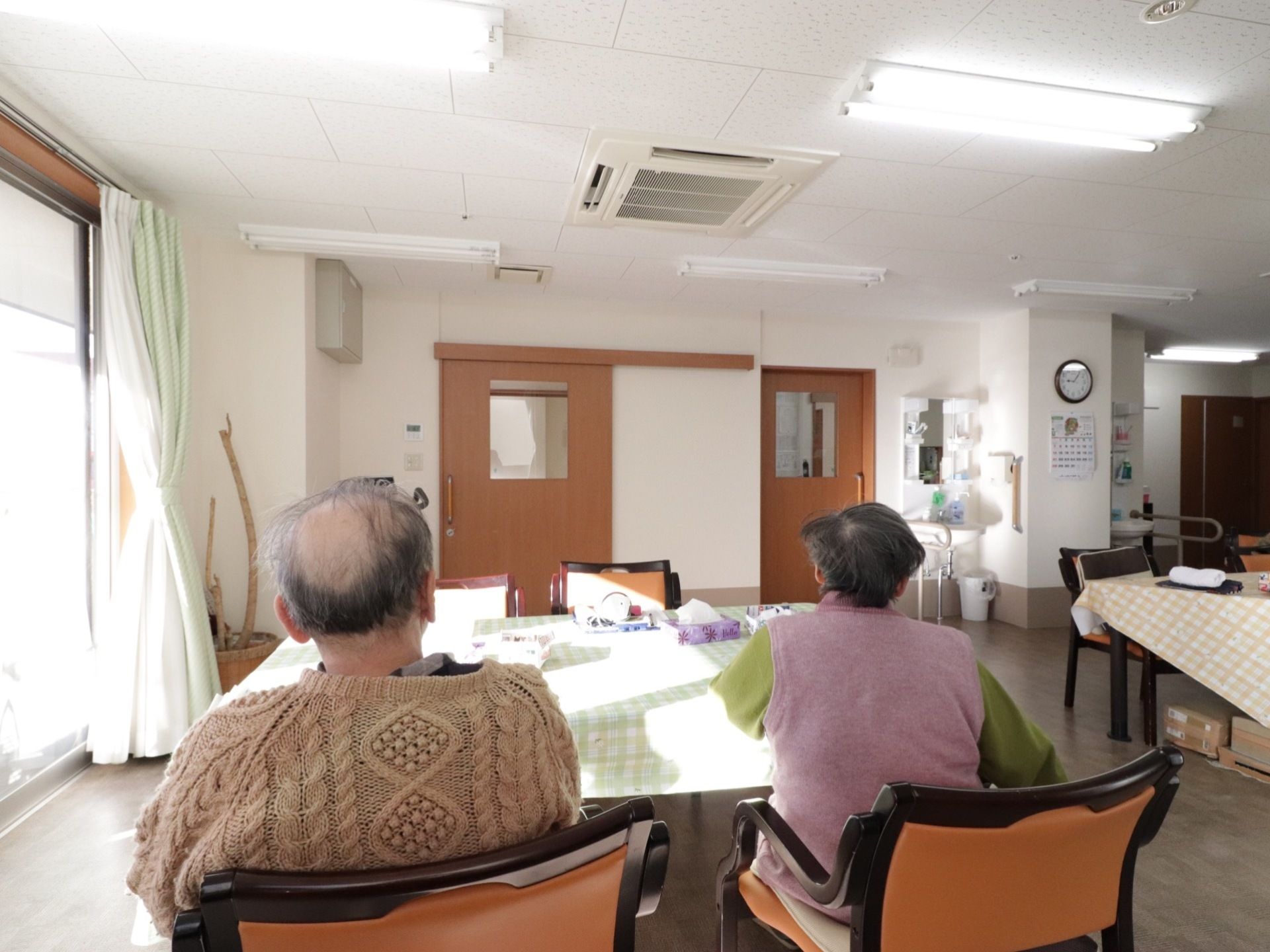 リビングで男女の入居者様が隣に並んで椅子に腰掛けている。テーブルの上にはティッシュ箱が置いてある。窓からは太陽の光が差し込んでいる。