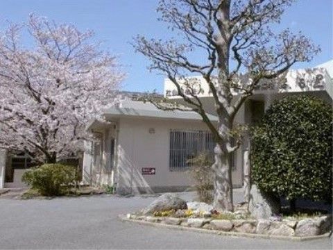 施設の隣にある介護老人保健施設である。白い壁の施設の前に大きな満開の桜の木があり、手前にも木や花が植えられている。