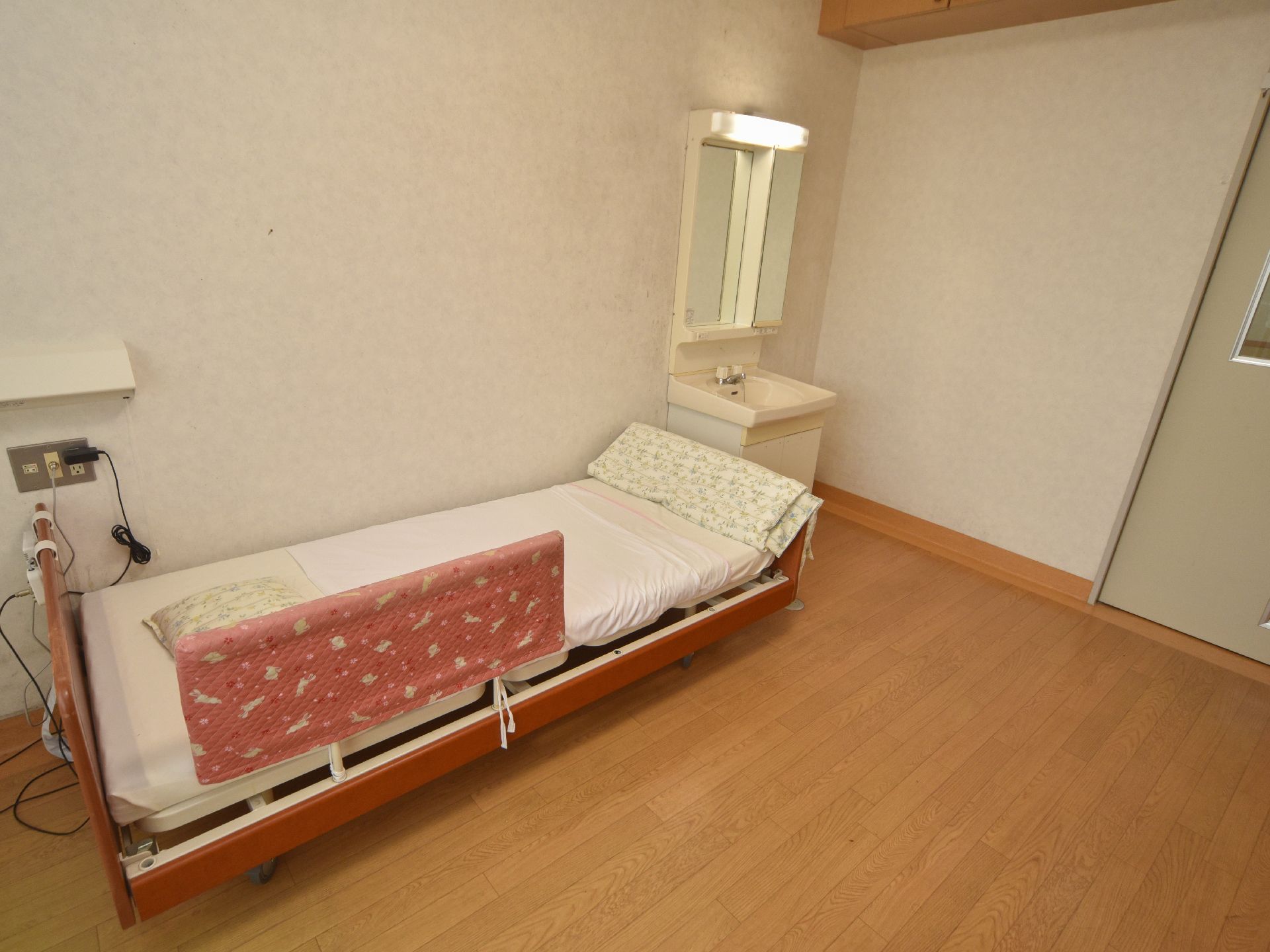 介護ベッドの枕元にはコンセントがあり、上には電気が付いている。ベッドの足元側に洗面台が設置されている。