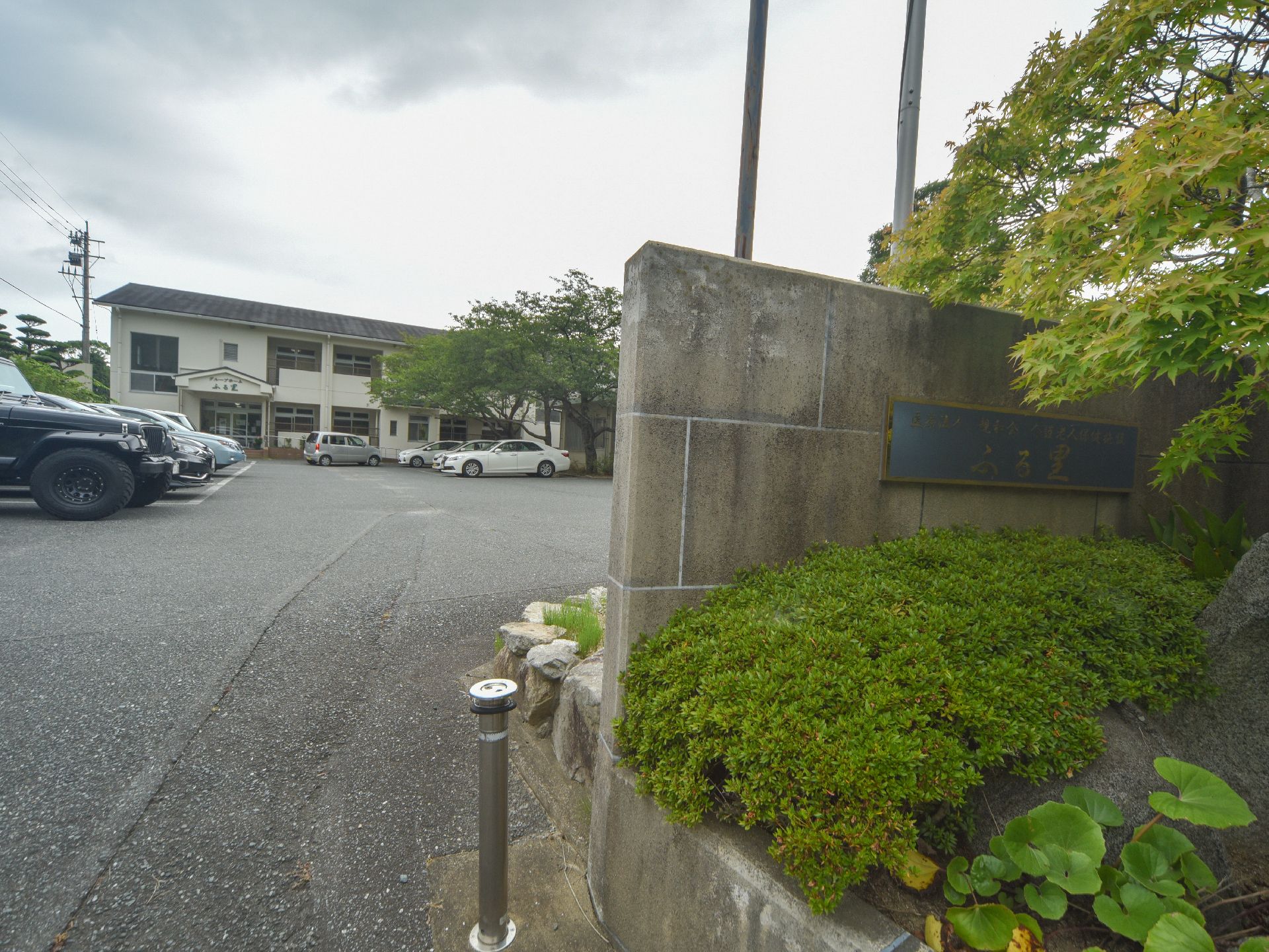 広い駐車場の向こうに建物が建っている。駐車場の外の塀に施設名が掲げられており、手前に紅葉やツツジが植わっている。