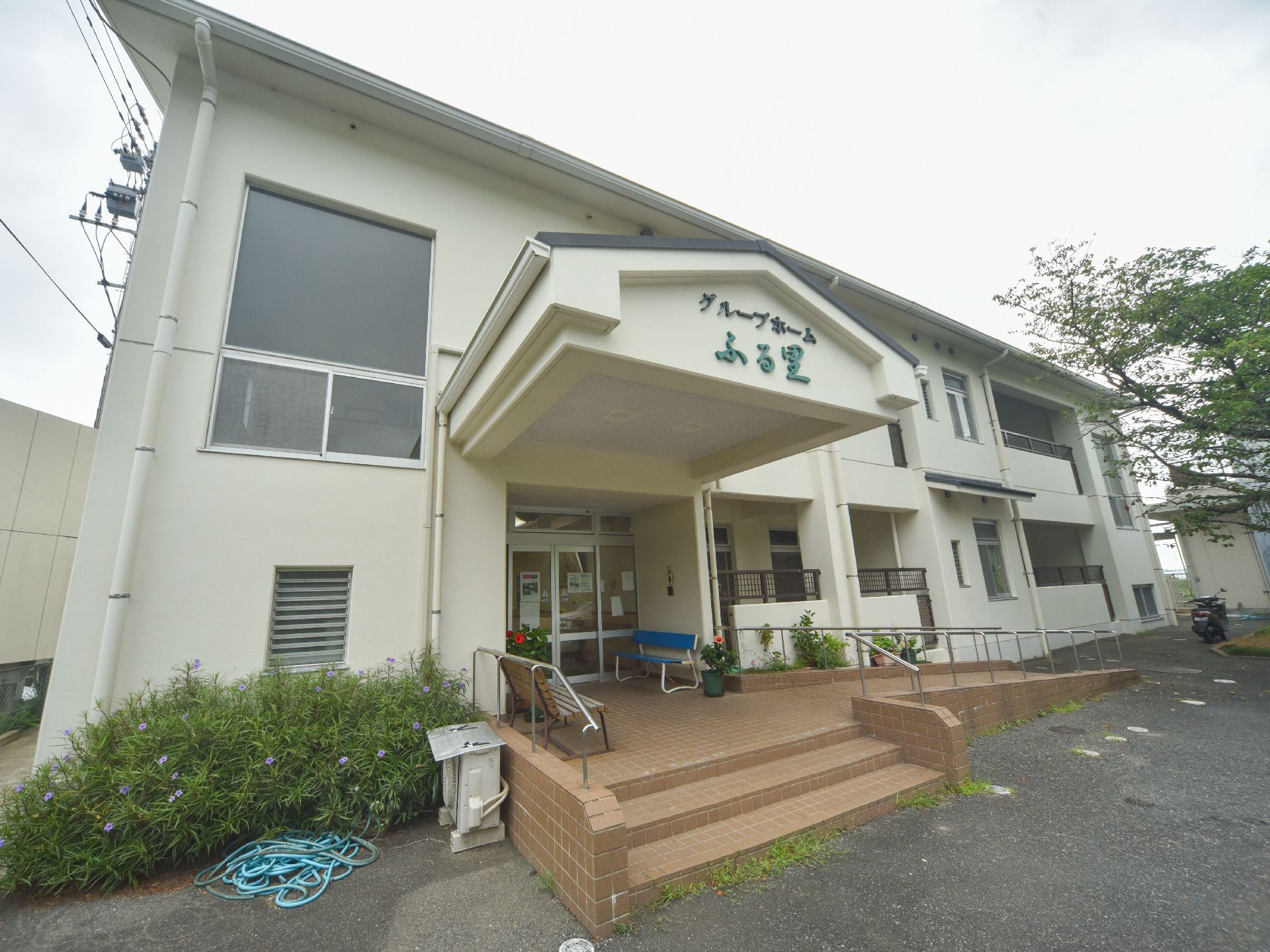 ポーチに木製のベンチとブルーのベンチが置かれている。建物は白い壁で、ポーチは茶色のタイル張りである。