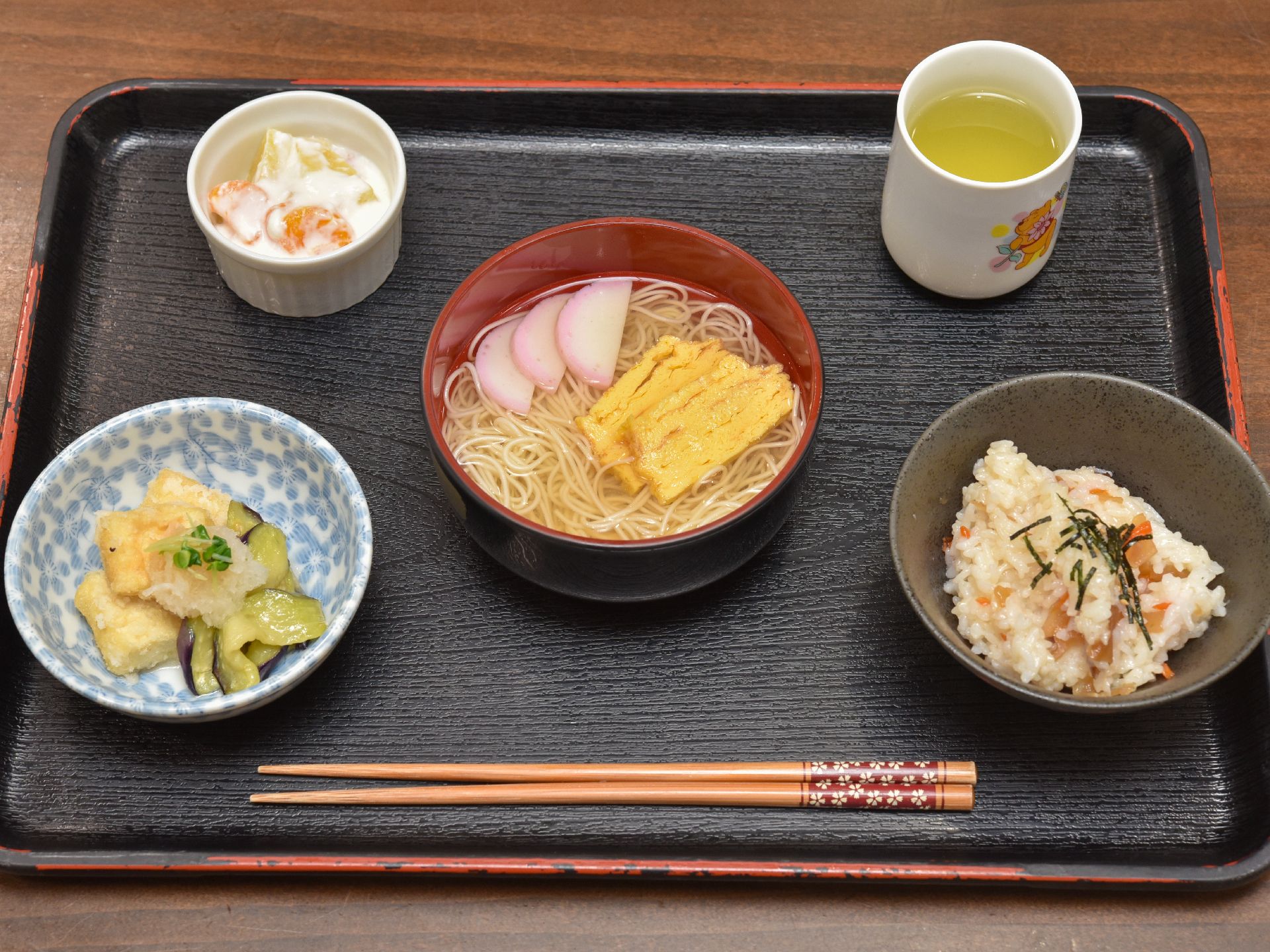 黒いトレーに食事と箸が載っている。どれも柔らかく食べやすそうなメニューで、デザートやお茶も付いている。