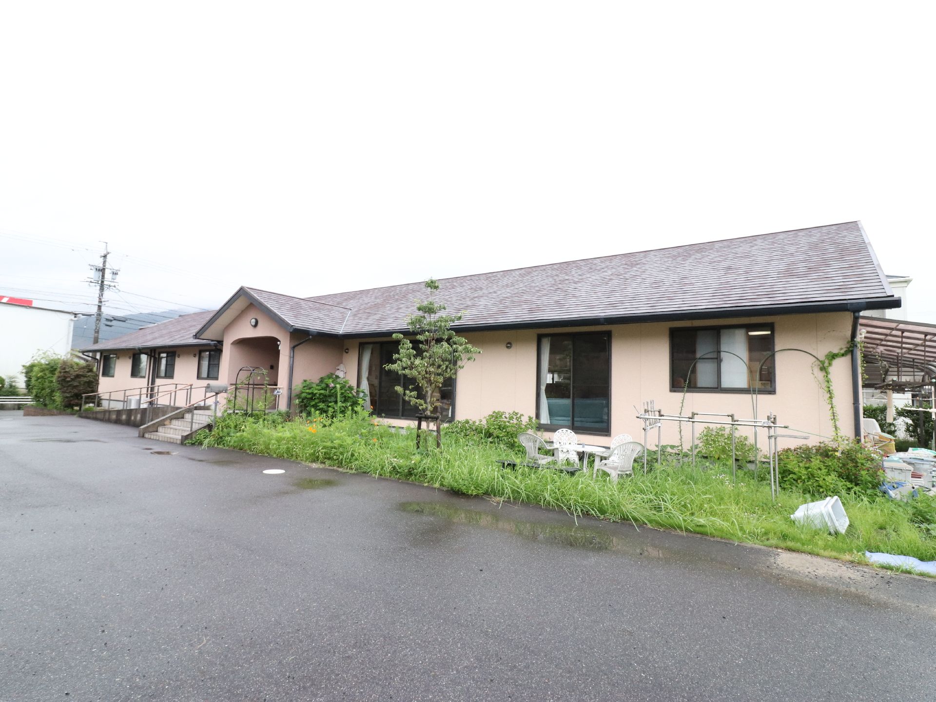 平屋建てで、前側には草木が植えられている。白いガーデンテーブルとチェアが置かれている。入り口は階段とスロープがある。