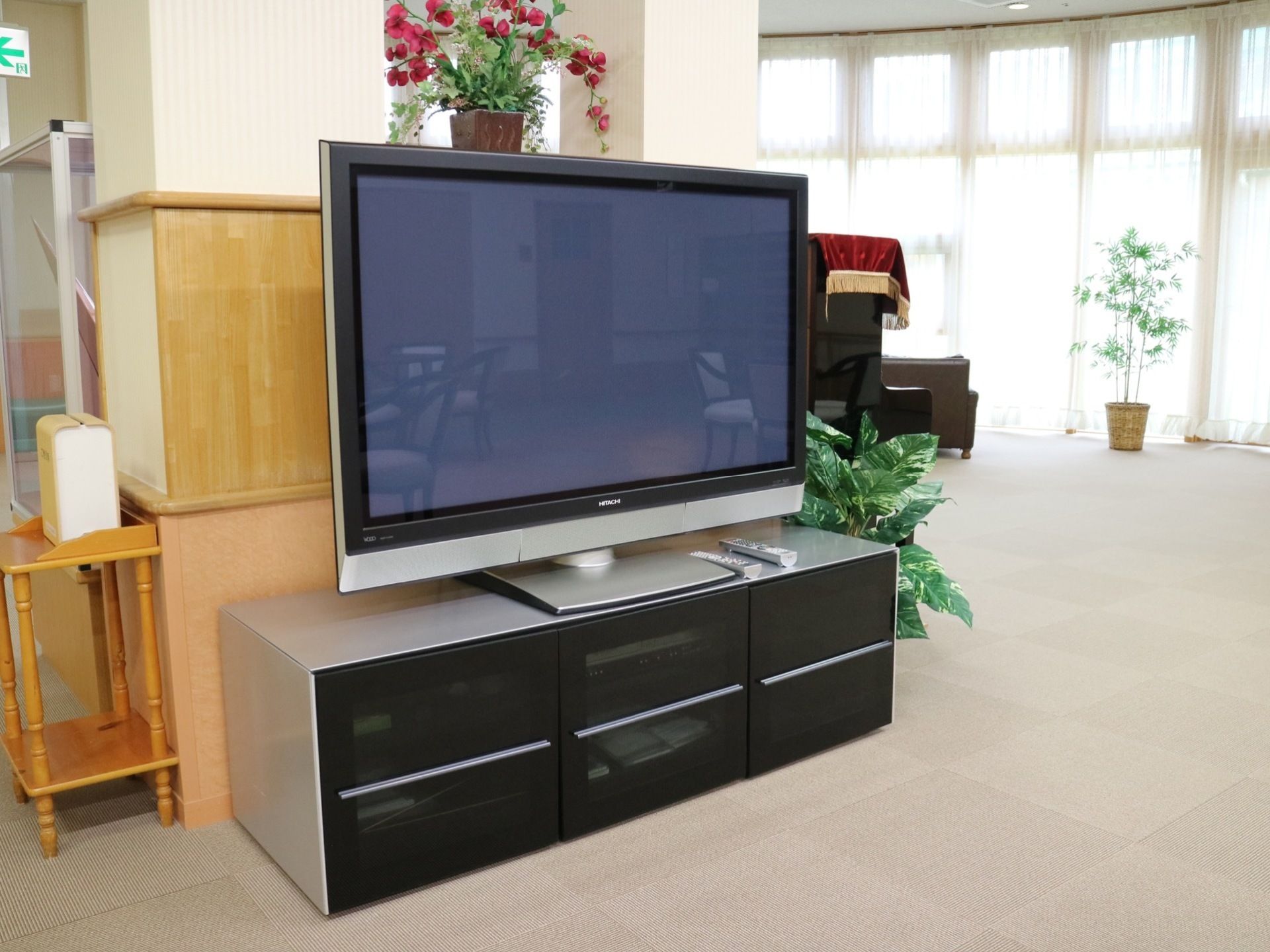 淡いグレーのフロアマットが敷き詰められた共有スペースである。室内には大きな液晶テレビや観葉植物などが置かれている。