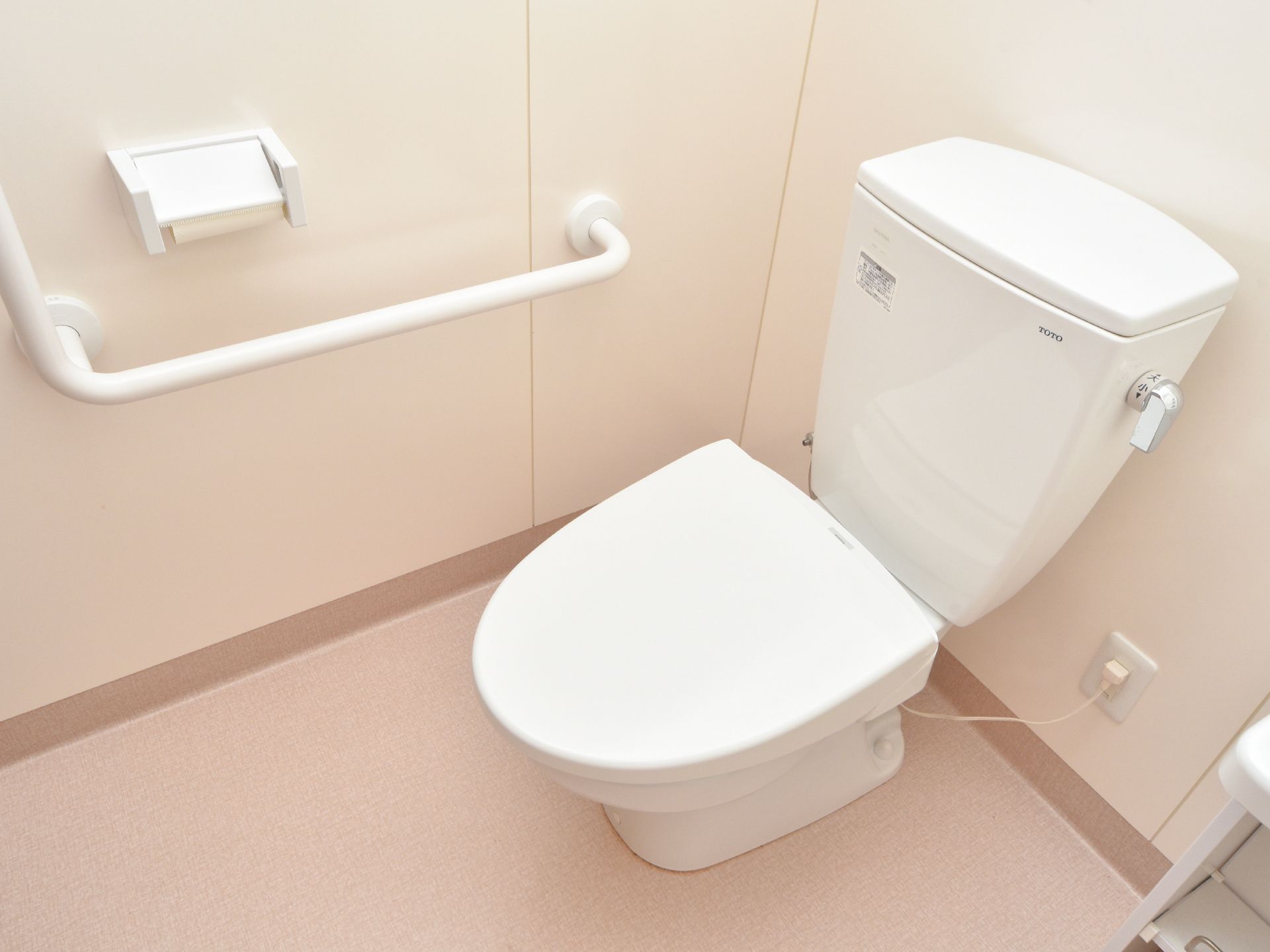 洋式トイレで、サイドの壁に手すりが付いている。トイレ本体、手すり、ペーパーホルダーがすべて白で、清潔感がある。