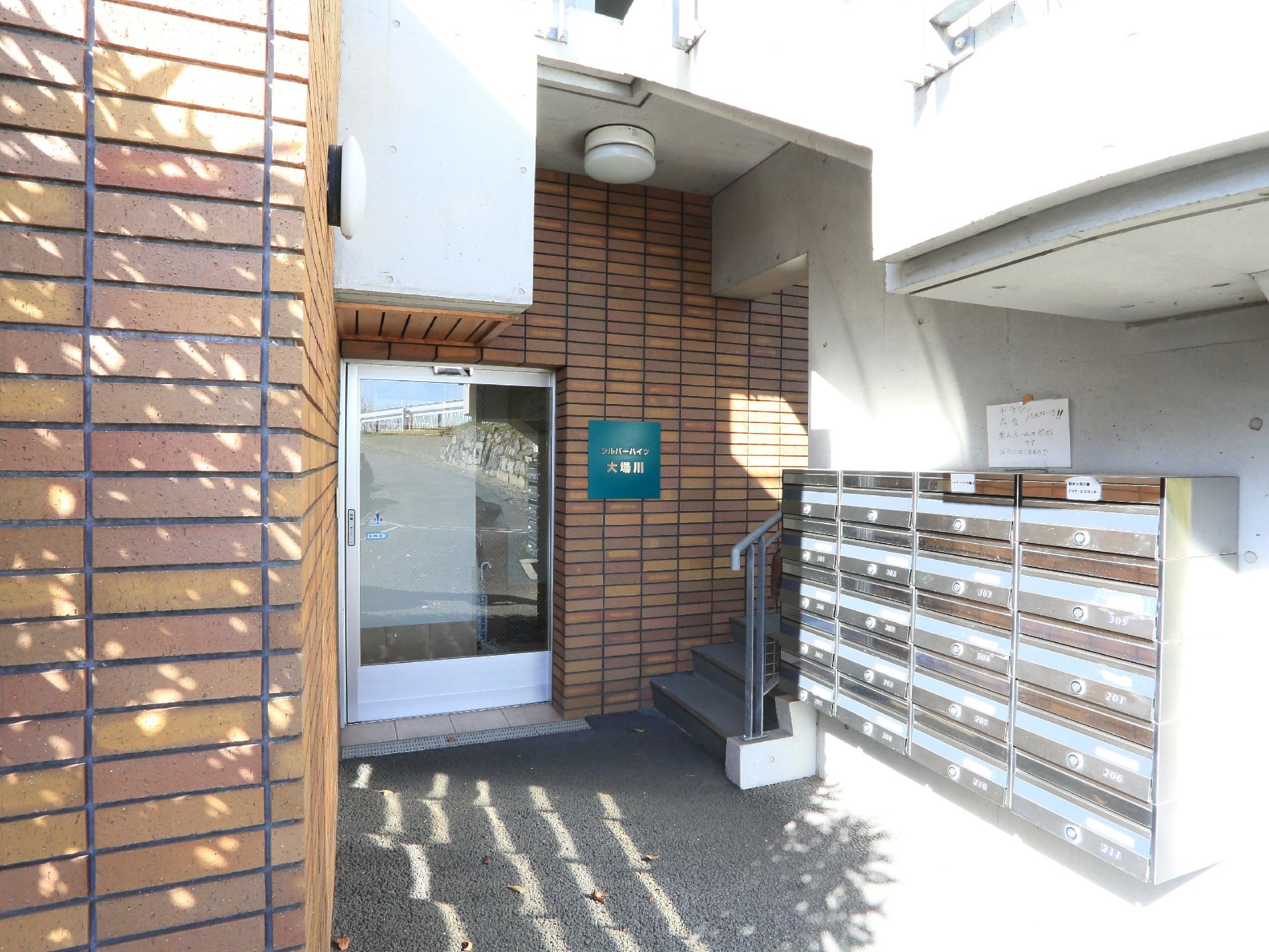 右側には沢山の郵便受けがある。正面には自動ドアがあり、その右側には階段がある。左側の壁とドアの上には照明が付いている。