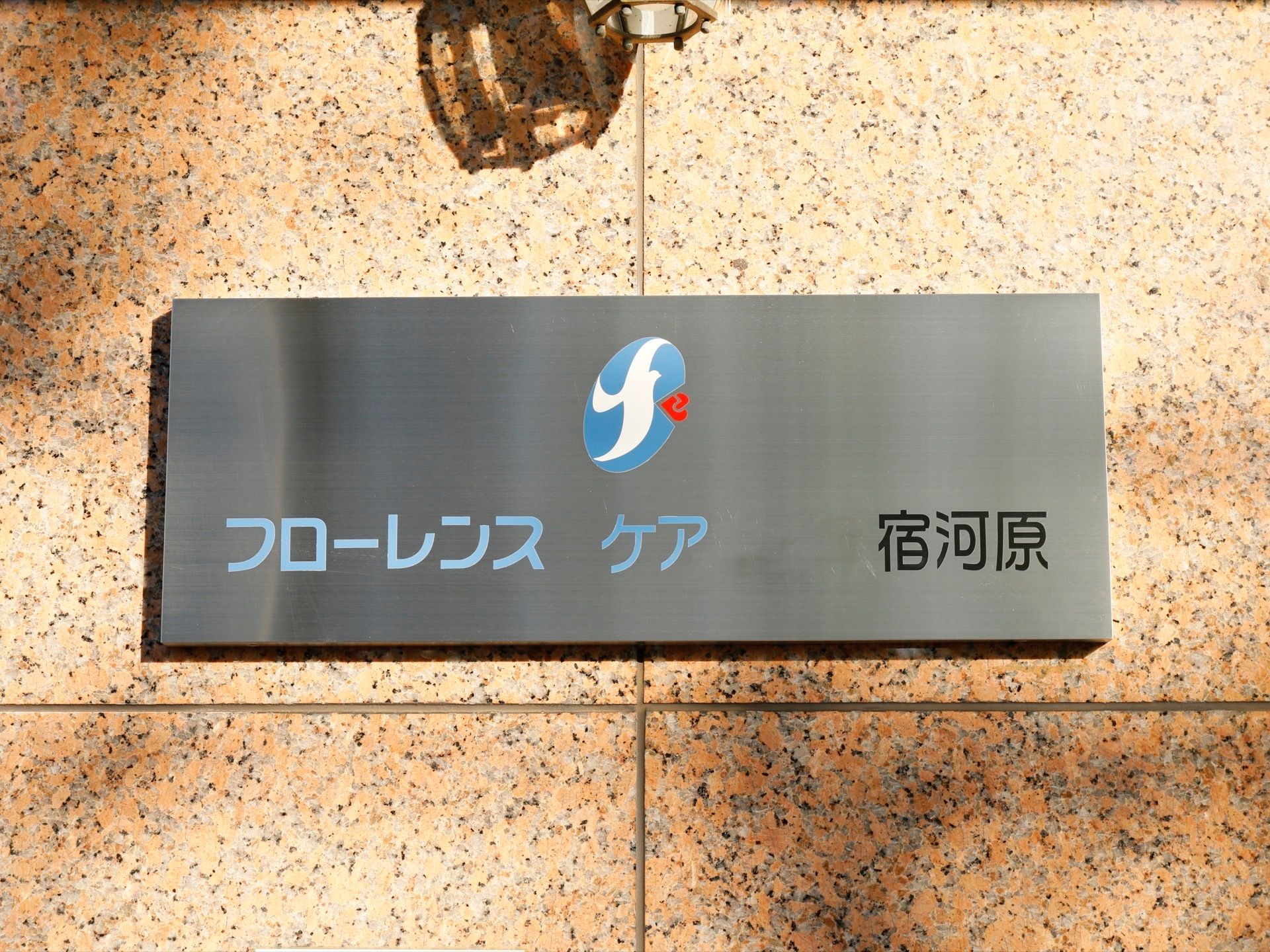 シルバーの板に施設名とロゴを表示した看板が建物壁面に設置されている。看板の上にライトがあるので、夜も見やすい。