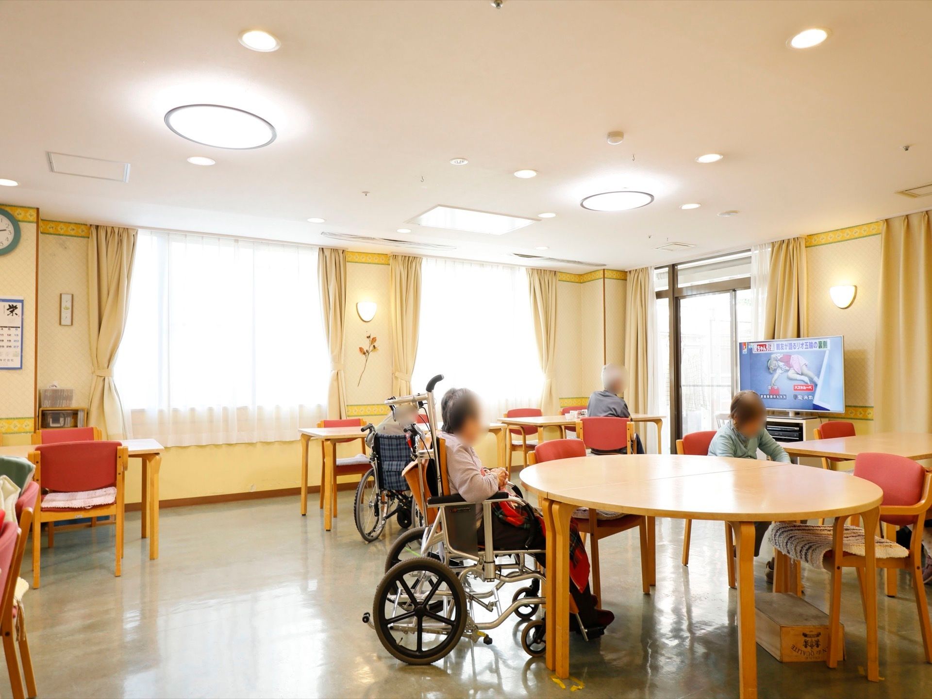 食堂に多くの椅子と、四角や丸の形のテーブルが置かれている。壁近くにはテレビがある。大きな窓や照明で明るい。