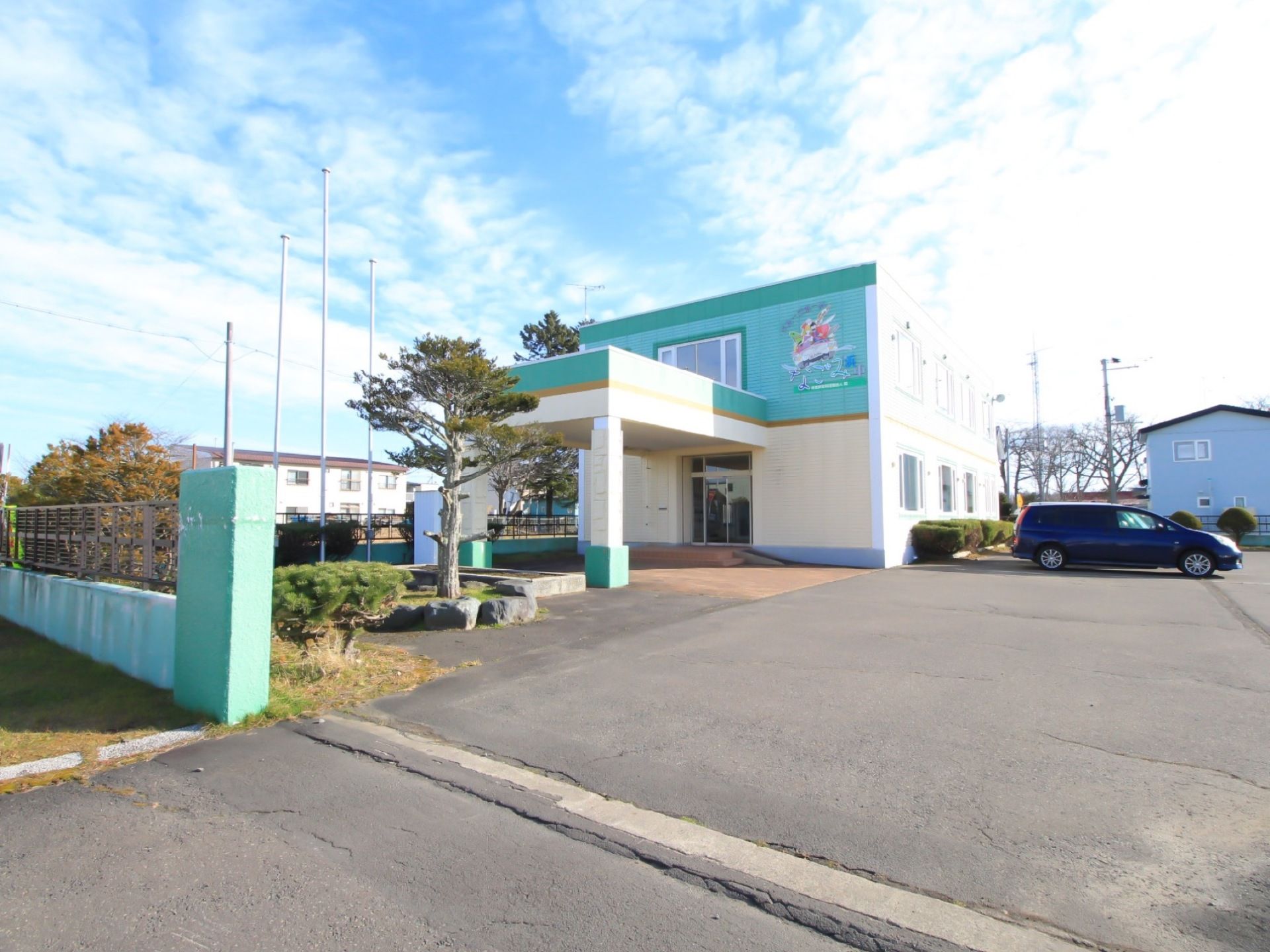 アスファルトで整備された敷地に車が停められている。緑色と白色のツートンカラーの２階建ての建物が建っている。