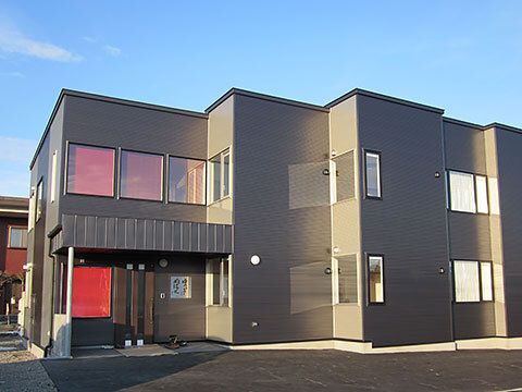 ダークグレーの外壁の２階建ての建物。敷地内には広い駐車場があり、日当たりのいい場所に建っている。