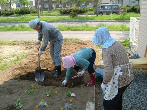 長袖長ズボンにエプロンや婦人用の農作業帽子を身につけ、畑で花を植えている女性たちと穴をスコップでほっている男性。