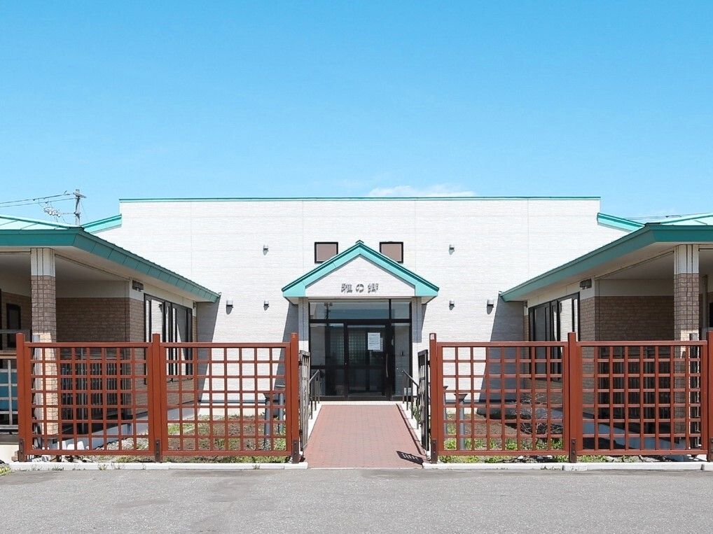 青い空の下には白を基調とした建物が建っていて、緑の屋根が付いている。建物の周辺には茶色いフェンスがあり、玄関前にはまっすぐな道がある。