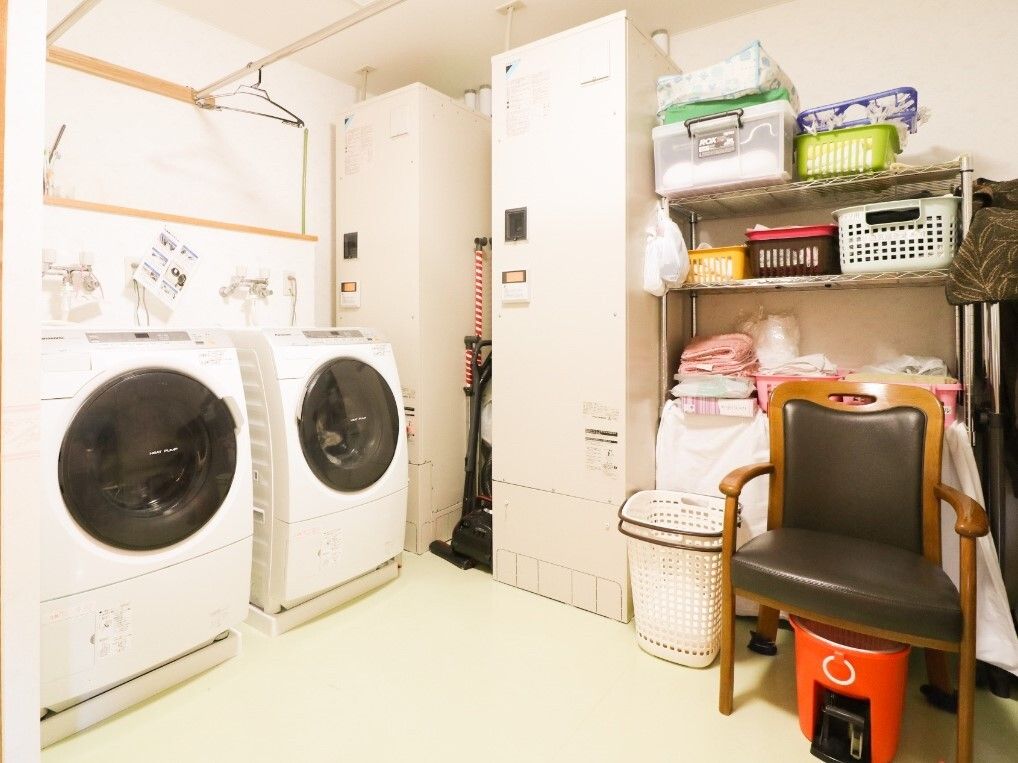 二つのドラム式の洗濯乾燥機が設置された洗濯室。棚にはタオルやプラスチックケースなどが並べられ、一人用の椅子も置かれている。