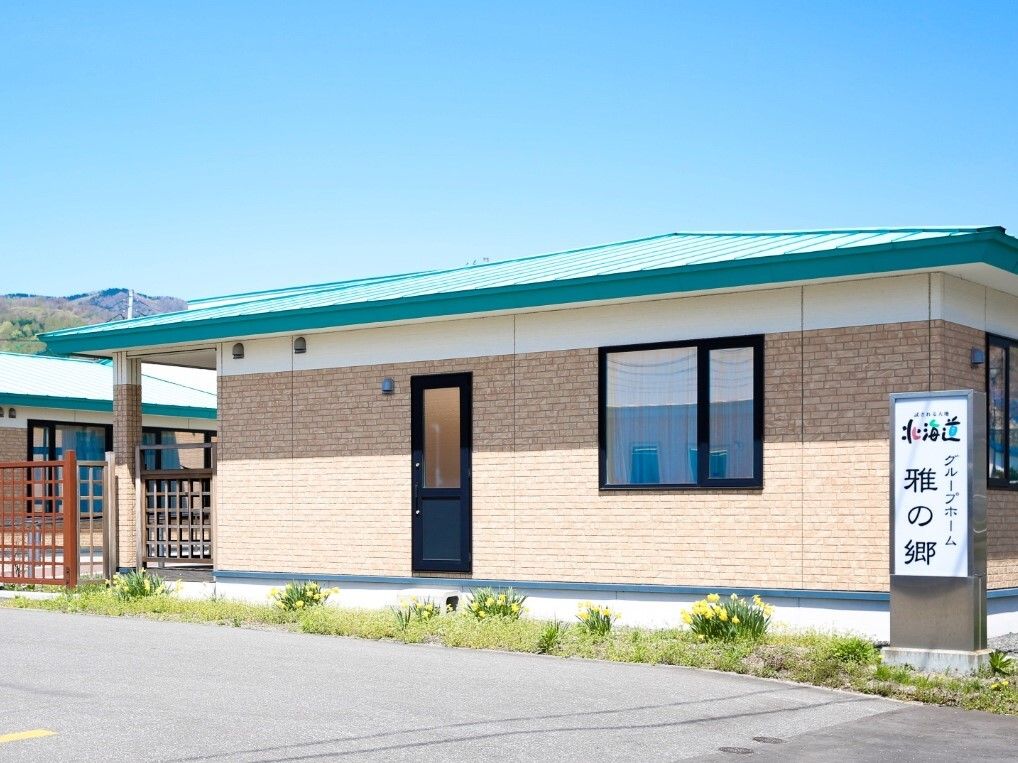 広い駐車場が付いた平屋建ての建物。ベージュ系の外壁にグリーンの屋根が付き敷地への出入り口にはホーム名が書かれた館銘板もある。