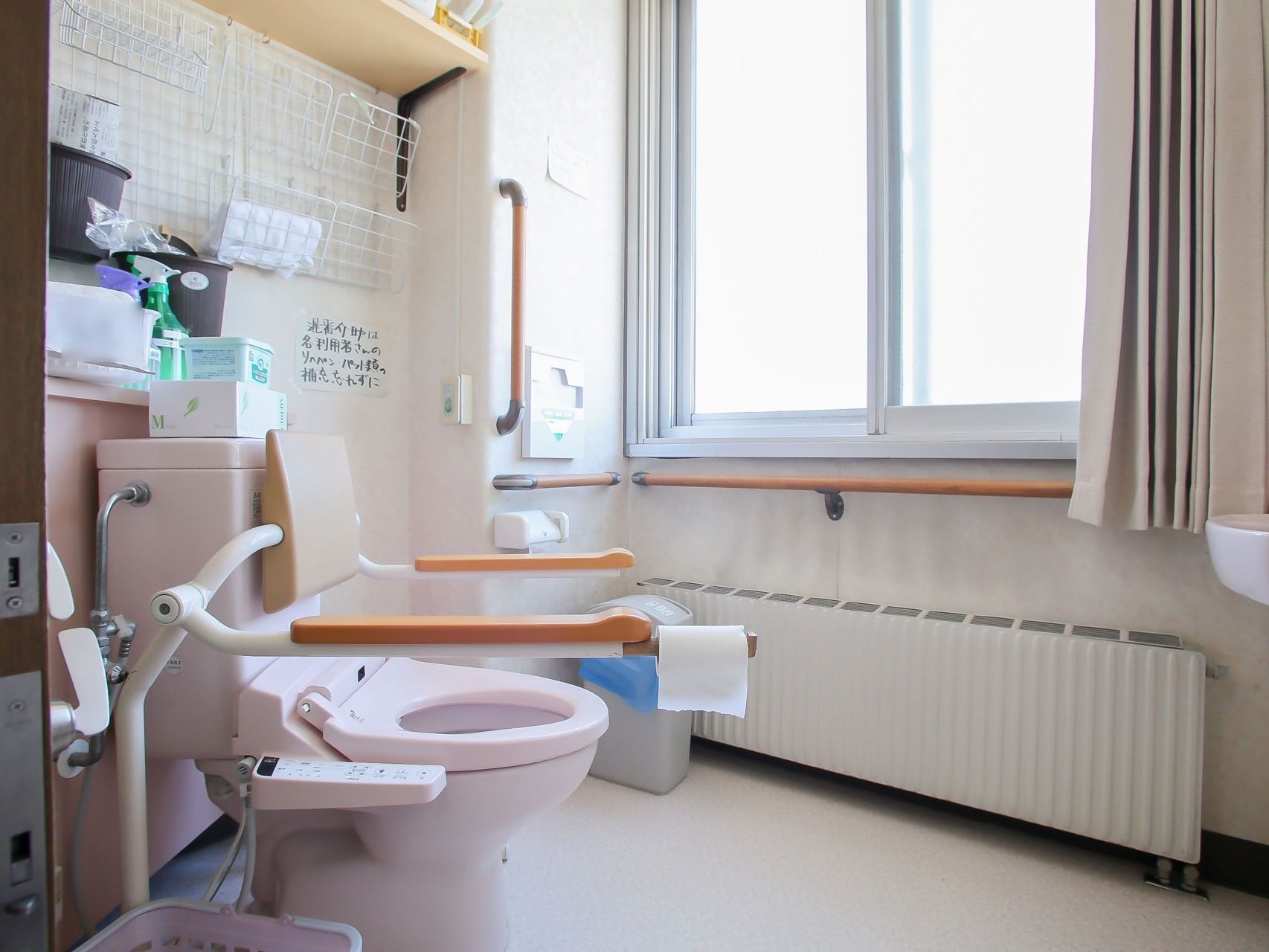 白で統一されたトイレは非常に清潔感がある。温水洗浄便座が設置され、背もたれと手すりがついている。清掃用具などがたくさん置かれている。