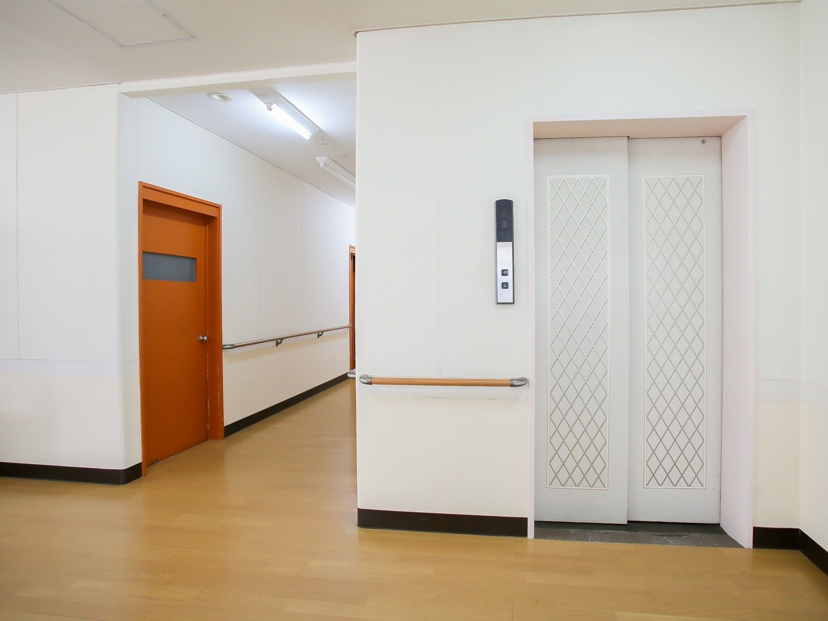 通路脇に設置されたエレベーターである。白い扉で壁に同調したデザインとなっている。そばの壁沿いには手すりが設けられている。