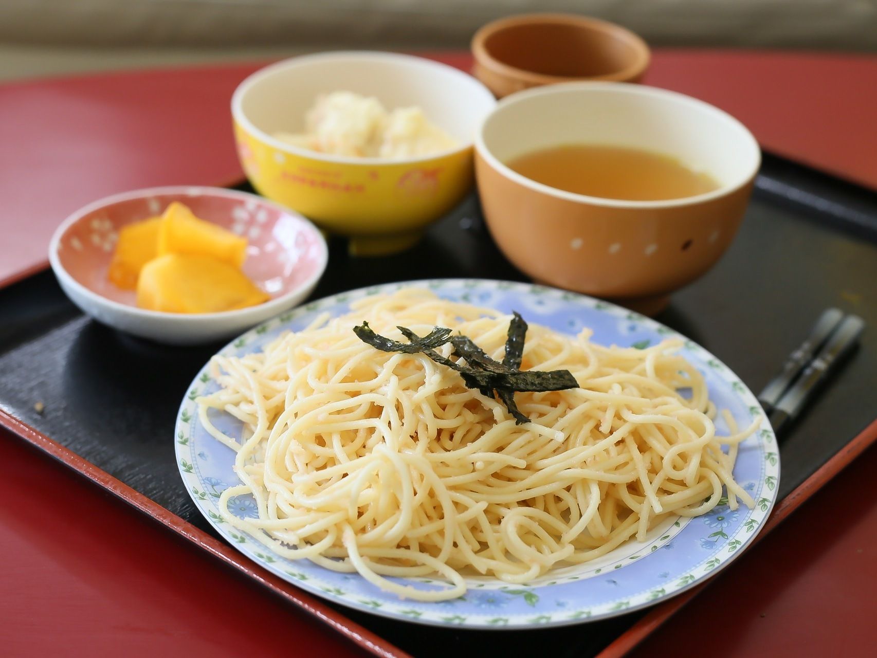 施設で提供される食事である。木製の四角いお盆の上にパスタをメインとしたメニューが盛られている。そばには箸が置かれている。
