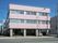 白い柱にピンクの壁が特徴の３階建ての建物が当施設である。玄関脇には駐車スペースがあり、車が停まっている。