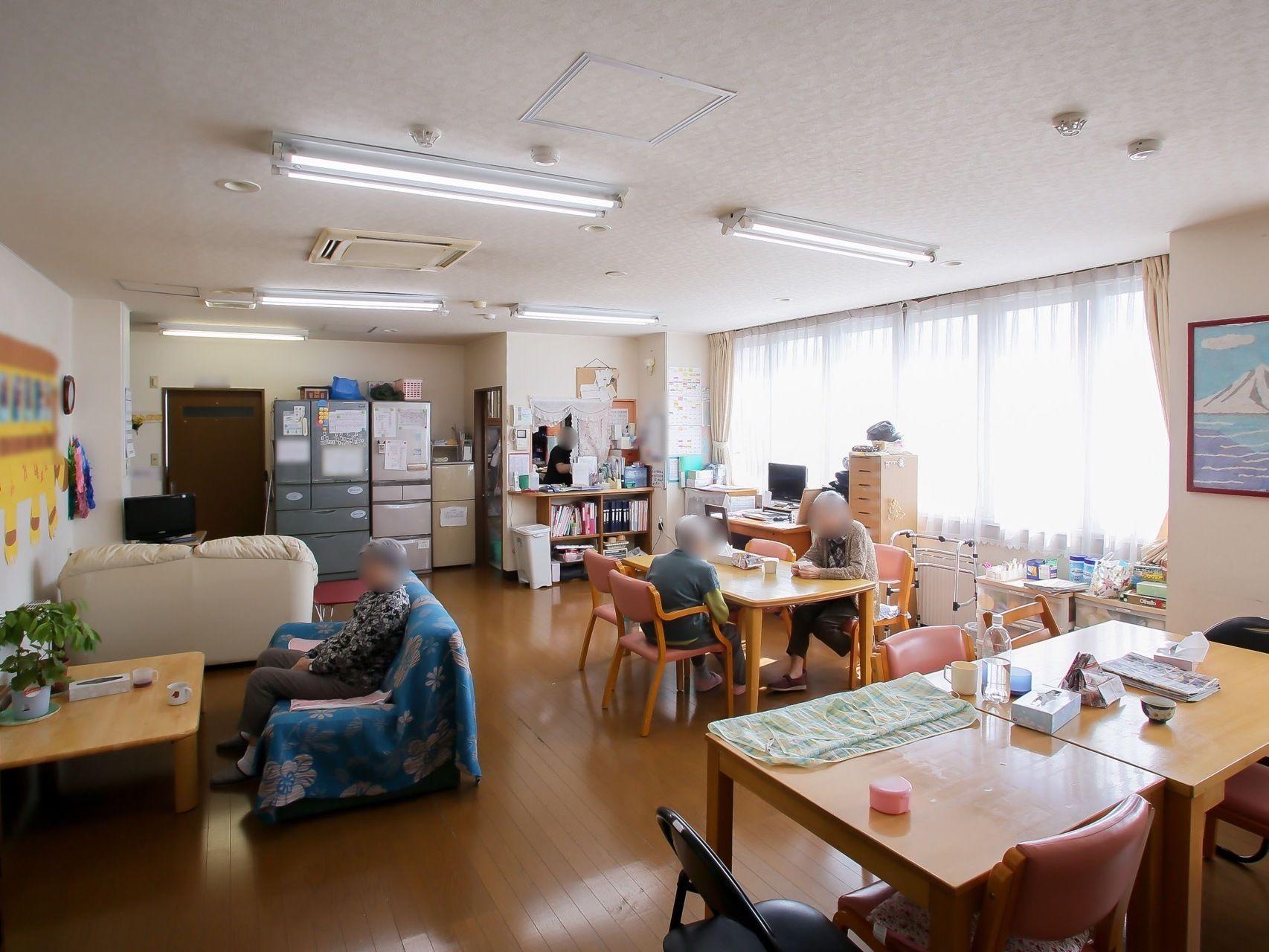 冷蔵庫と小さな台所のあるリビングで、ソファやテーブル席で時間を過ごす入居者様たち