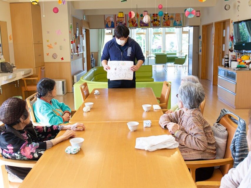 マスクをしたスタッフが入居者が座っているテーブルの前に立ち、何か紙のようなものを見せている。