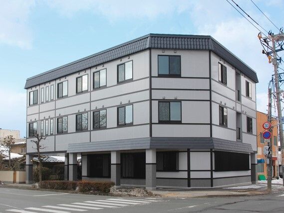道路の面して建っているグレー系の3階建ての建物の周りには、草木も植えられている。