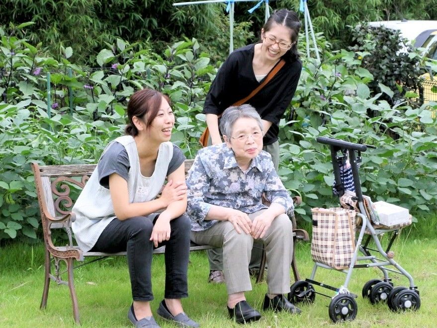 外で散策している様子の高齢者とスタッフの様子。荷物を置ける手押し車がある