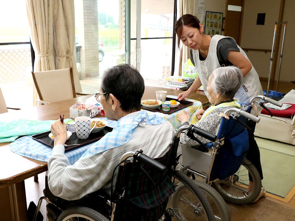 食事介助している様子を写したもの。車いすの高齢者とそれを見守るスタッフ