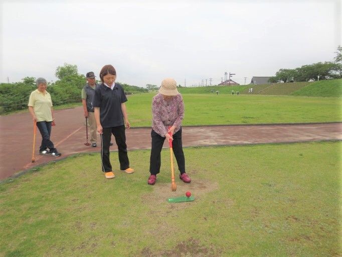 帽子をかぶった男女の入居者がグランドゴルフを楽しんでいる様子。スタッフが付き添っている。