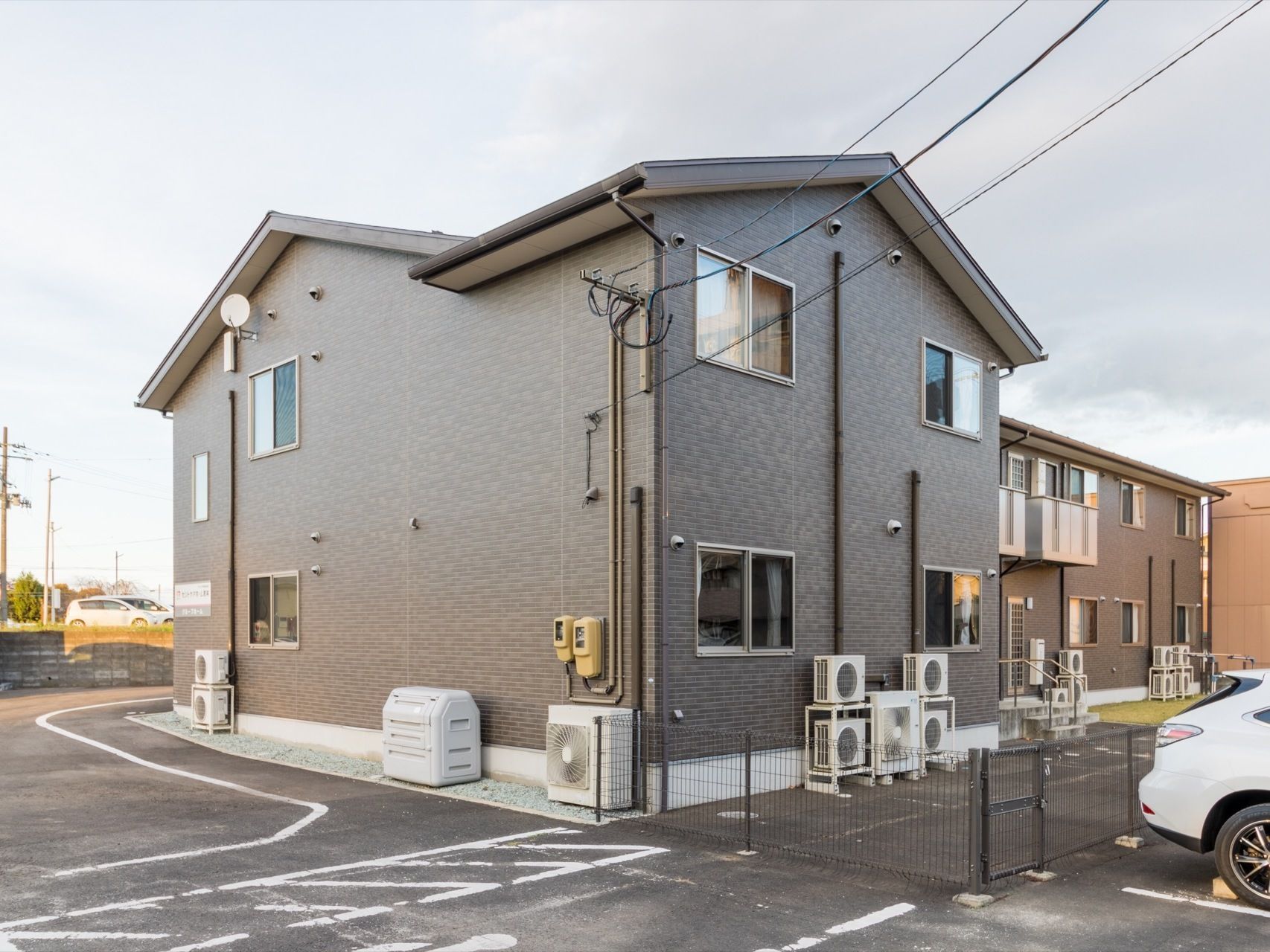 グレーのモダンな色合いの外壁である。２階建て一軒家のような見た目の建物である。敷地や周辺は広く、ゆったりとしている。