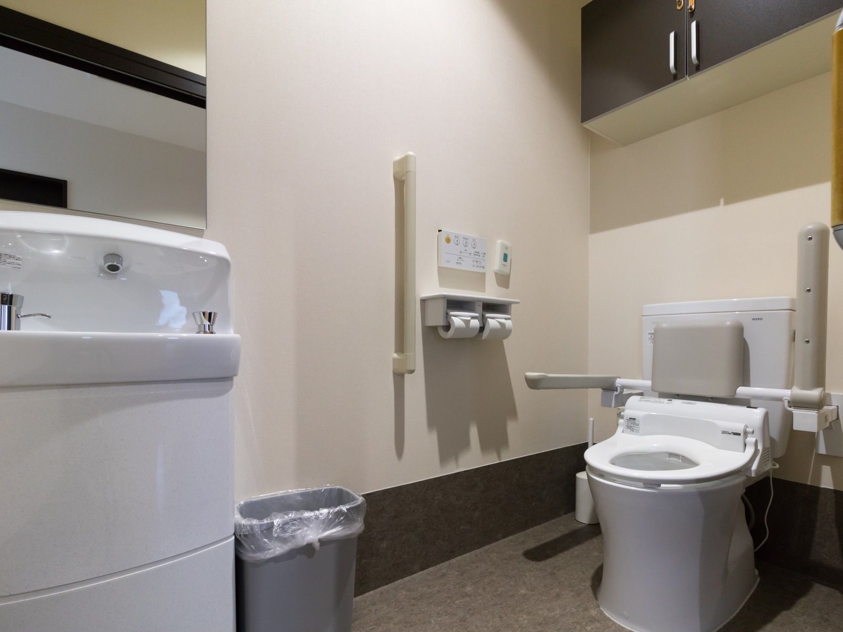 広く清潔な共用トイレである。便座は蓋を無くし、クッション性のある背もたれと肘掛けが取りつけられている。