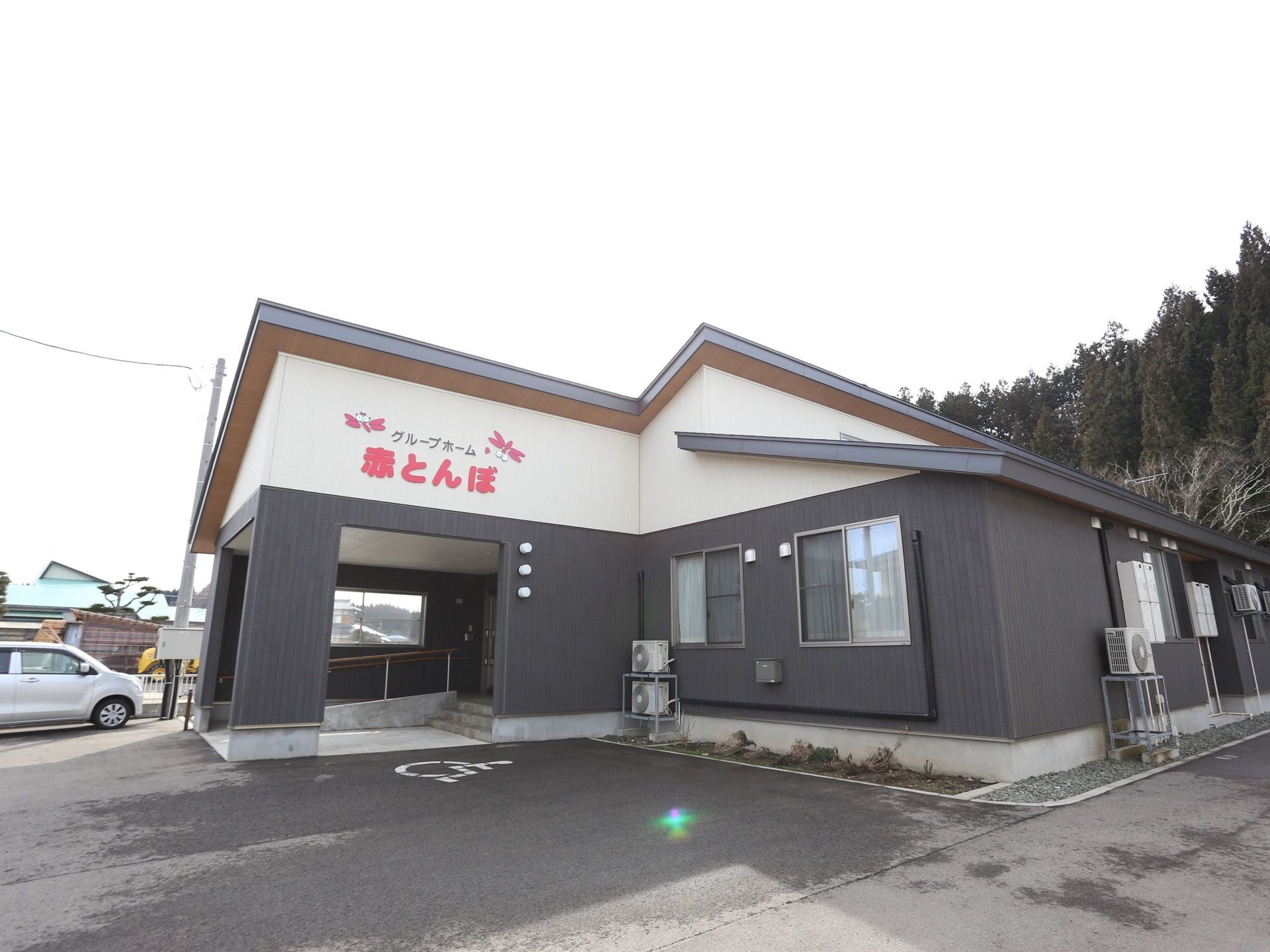 白とダークグレーで塗り分けられた外観の建物である。左手に玄関ポーチがあり、その前は車椅子専用車のスペースがある。
