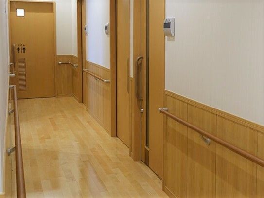 手すりが付いたまっすぐな廊下。奥にはトイレのマークが付いた扉があり、居室には木製の引き戸が付いている。