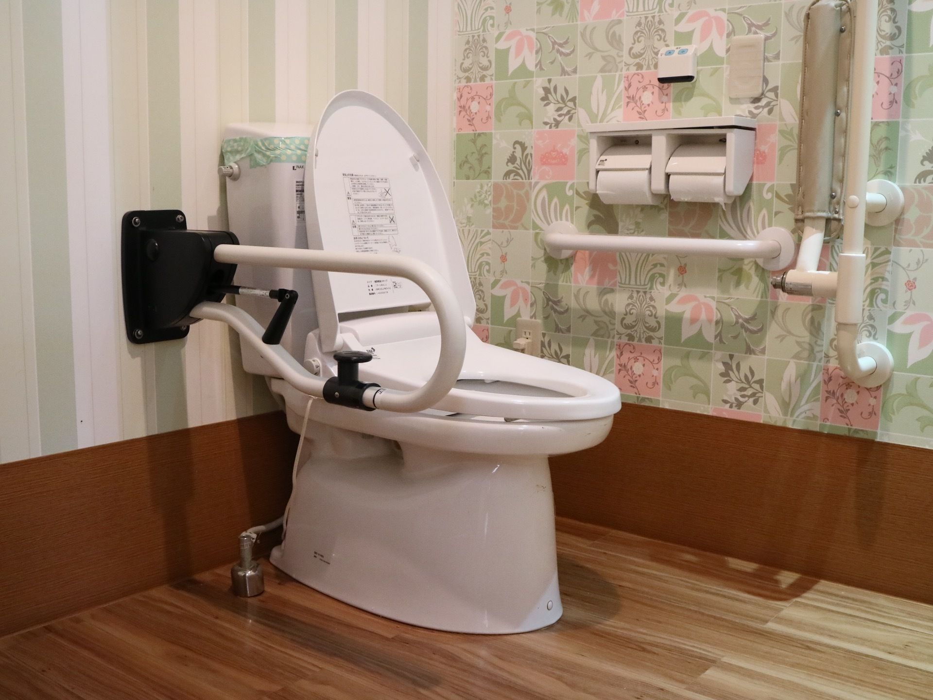 フローリングの床に、花柄や縦縞の壁紙のトイレである。中央に温水洗浄便座が設置された便座がある。壁に手すりがかかっている。
