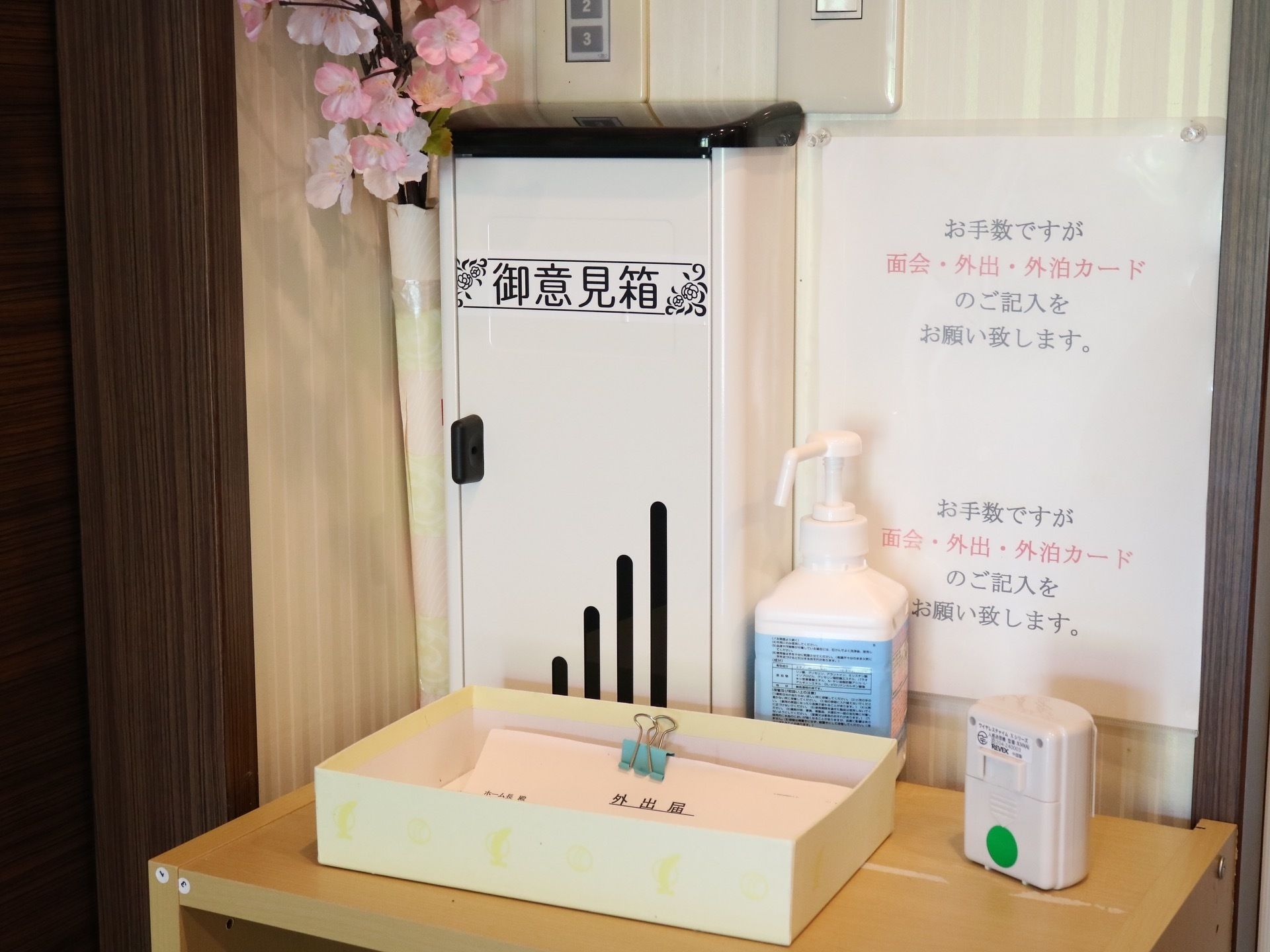 木製の棚に外出届け出用の用紙や意見箱、消毒用アルコールスプレーが置かれている。桜の花の造花が飾られている。
