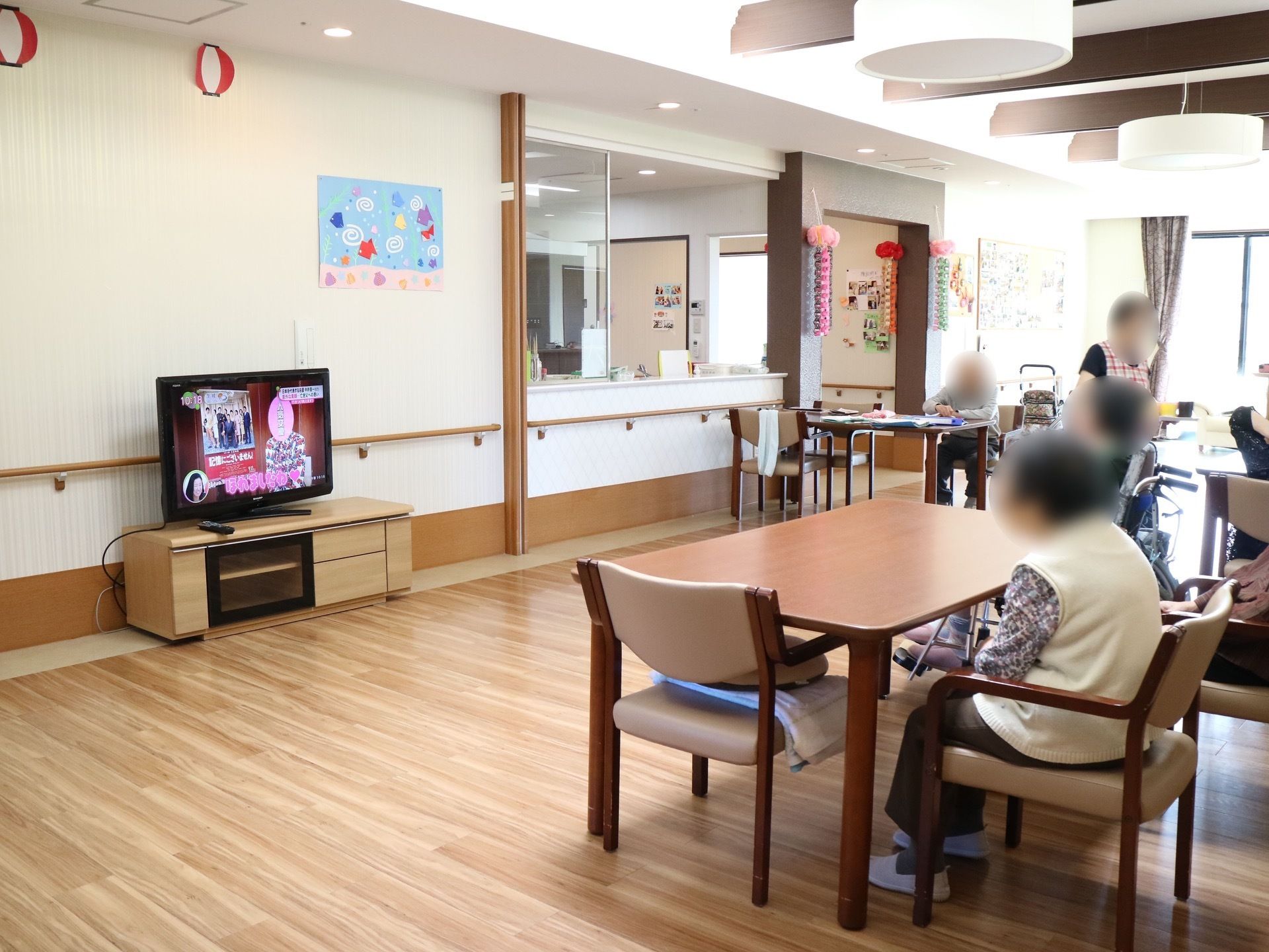 壁際に大型液晶テレビが置かれ、テーブルセットで入居者様が腰をかけて鑑賞を楽しんでいる。壁に飾り付けがされている。