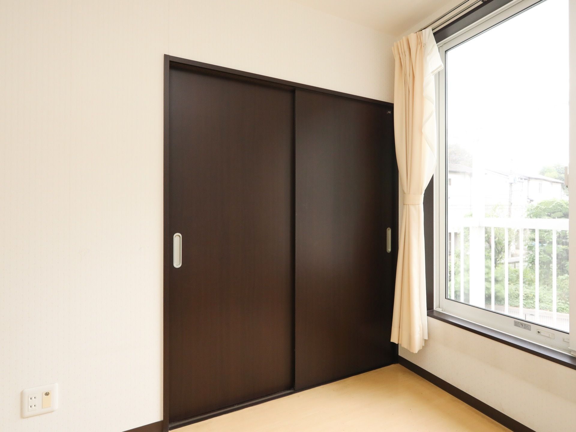 居室にはこげ茶色の引き戸の収納があり、扉の両端には銀色の取っ手が付いている。側には窓があり、カーテンが束ねられている。