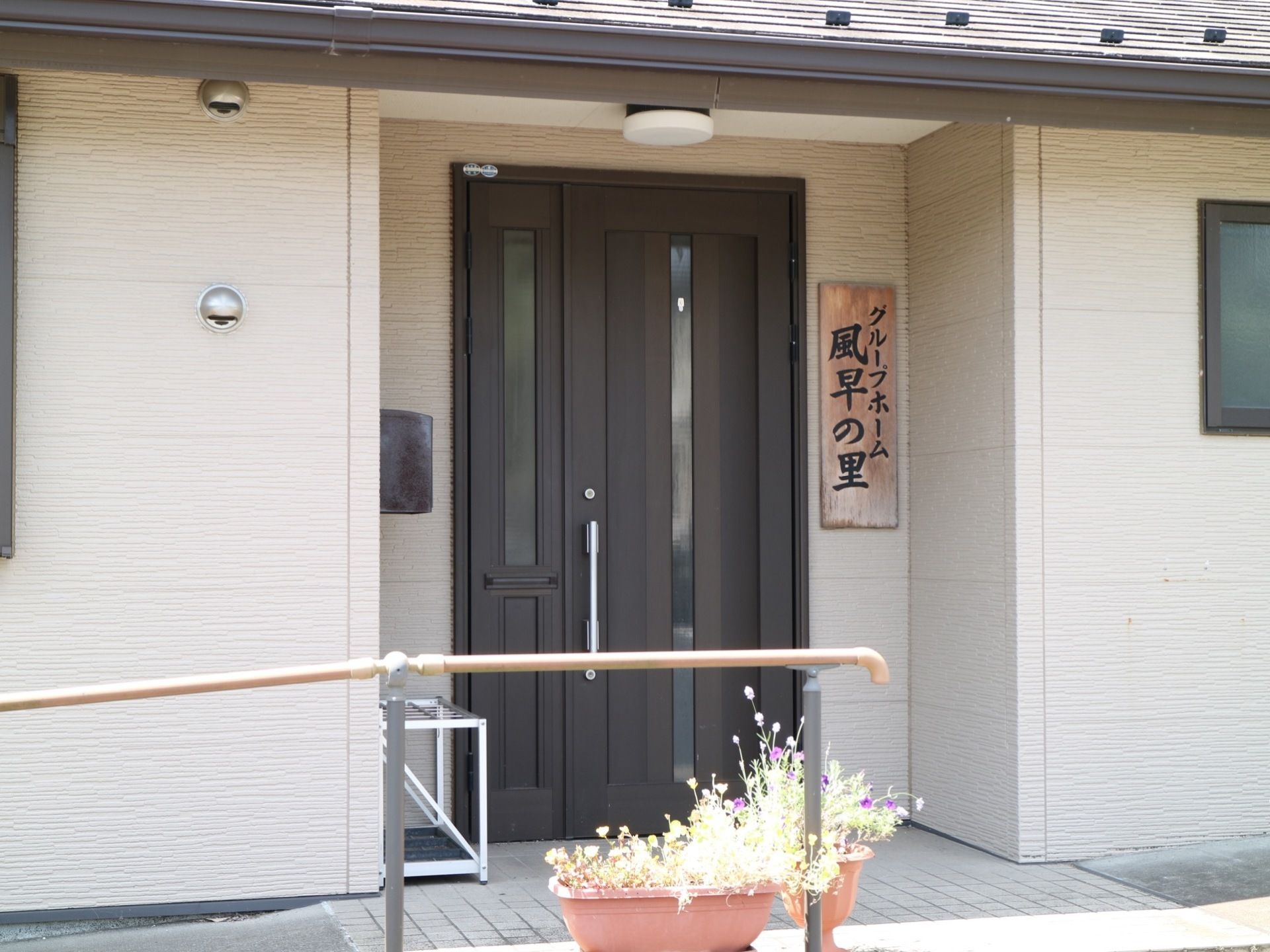 銀色の取っ手の付いた黒いドアがある。その横に木製の大きな表札があり、黒い文字で施設名が描かれている。
