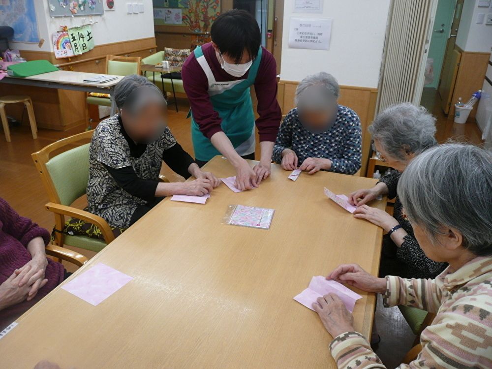 ベッドに寝ている高齢者女性に話しかけている女性スタッフは花柄のエプロンを付けている。