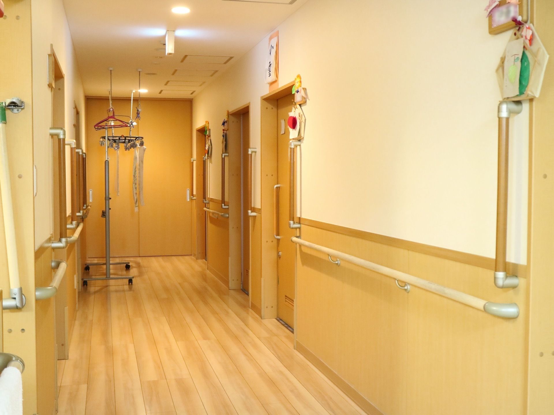 通路は白色と木目調のデザインである。壁沿いには水平方向だけでなく縦方向にも手すりが設置されており、居室のドアのそばに飾り付けがされている。