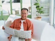 カメラ目線でほほ笑む男性。新聞を読みながらこちらを見ている様子。白いソファに座っている