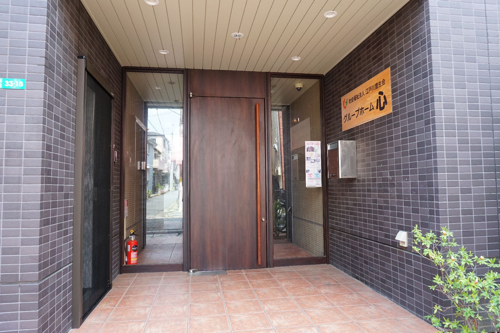 ブルーのエプロンを付けた女性スタッフに付き添われて屋外を散歩しているブラウンのジャケットを着ている高齢女性。