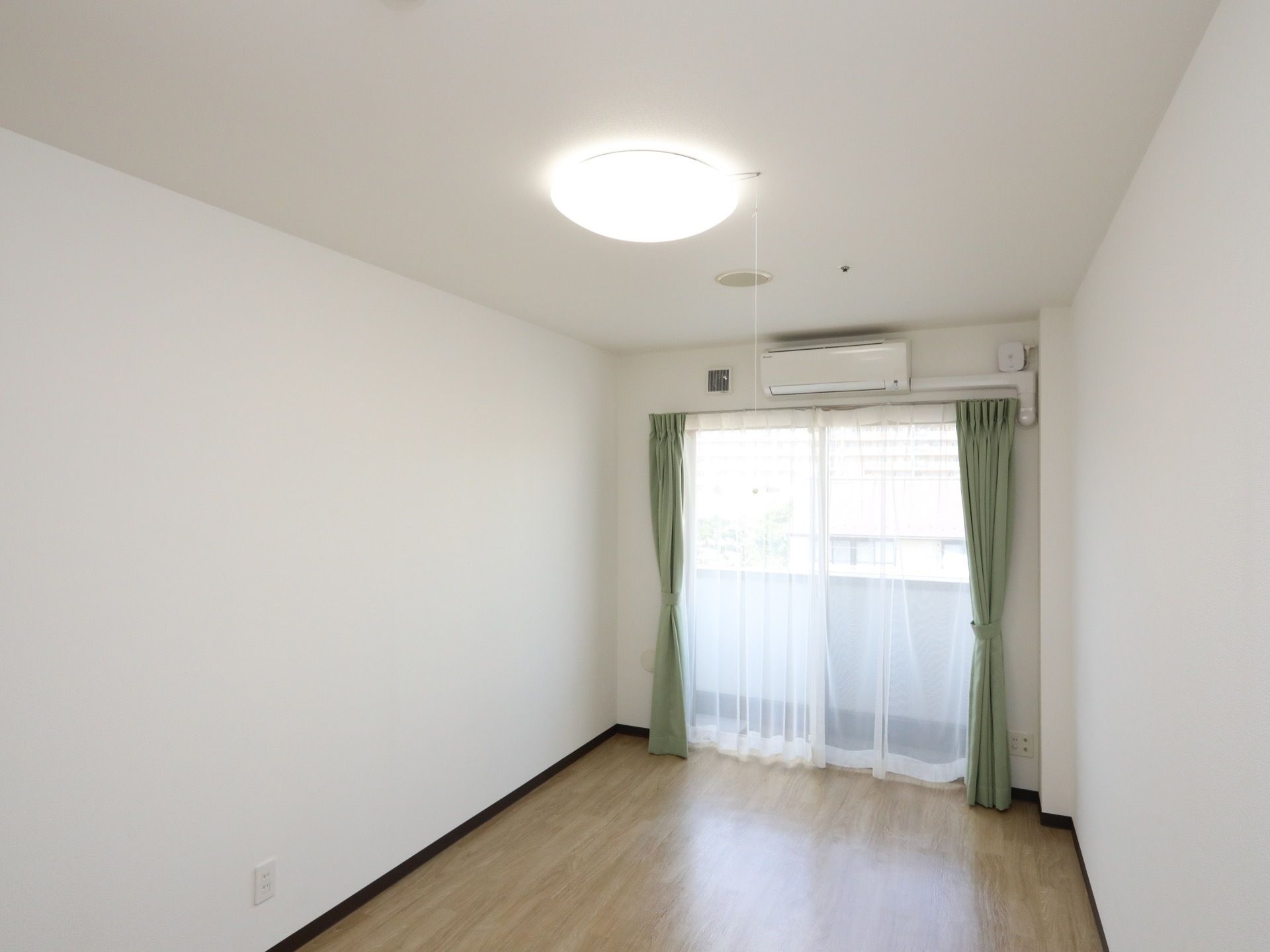 居室は奥行きがあり、大きな窓がある。床は全面フローリングで、天井には丸いシーリングライトが取りつけられている。