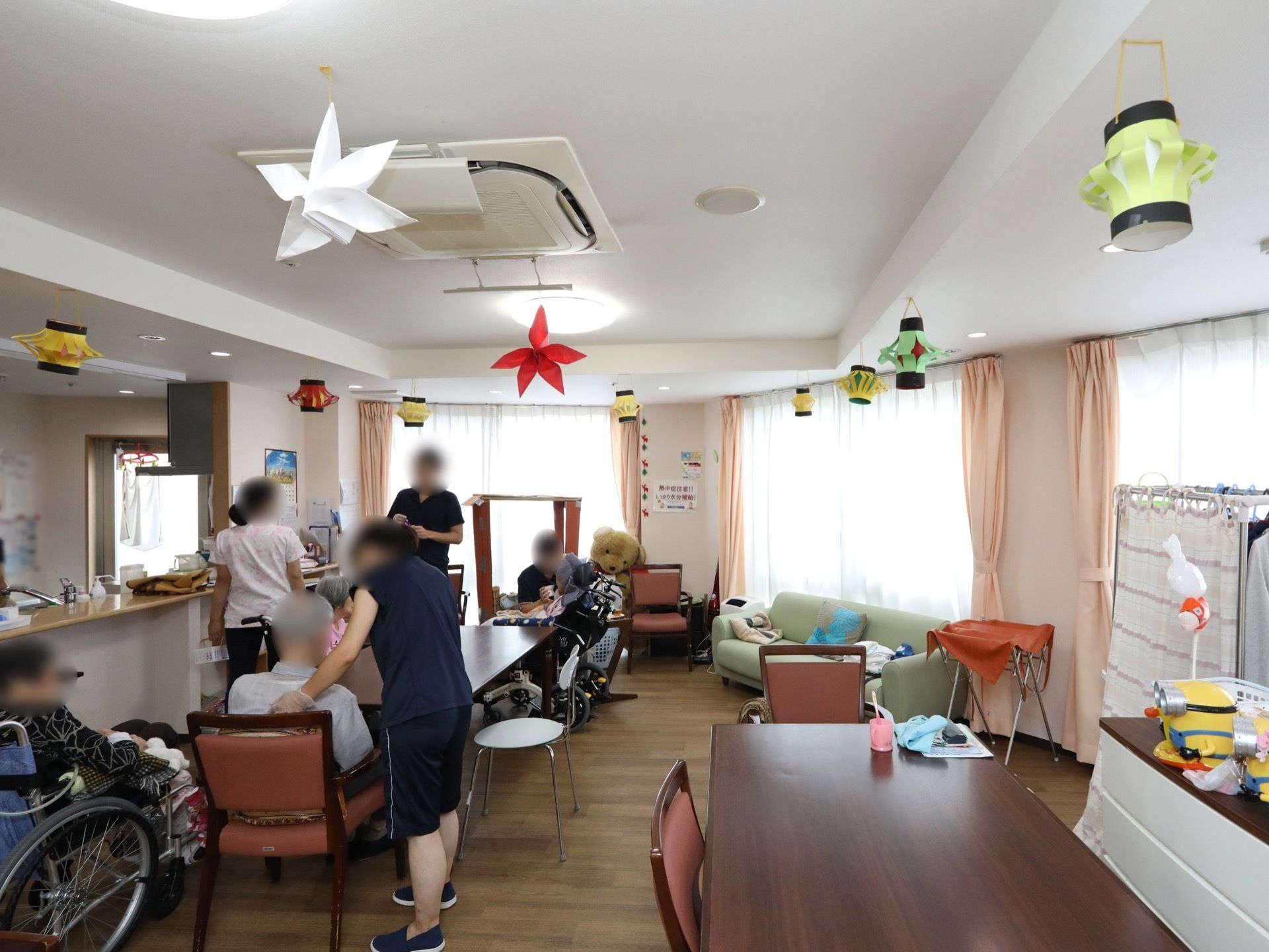 広々とした共有スペースには、大きなテーブルが２台置かれている。ソファーや車椅子に座った入居者様が、ゆったりと寛いでいる。
