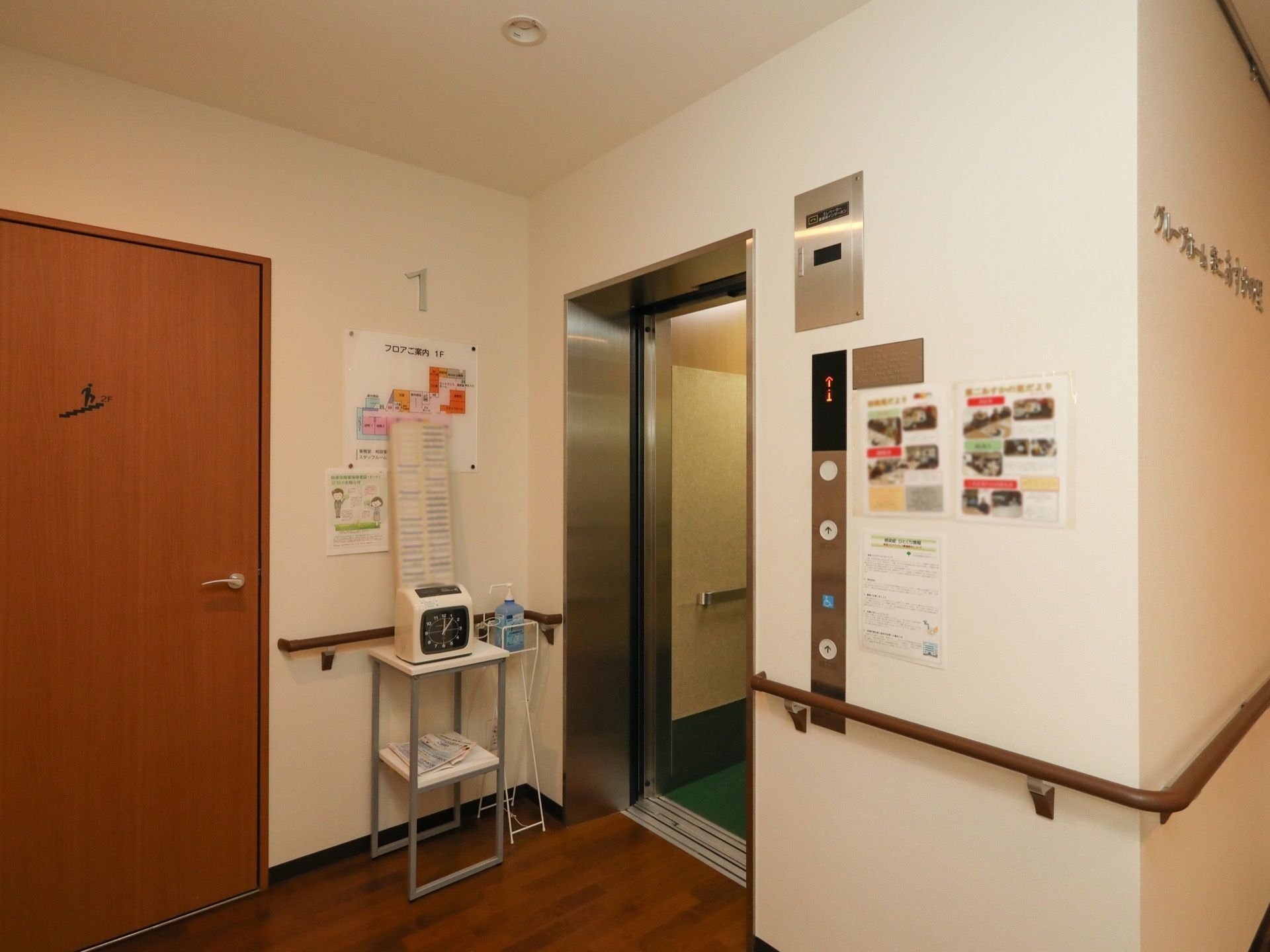 廊下の角にエレベーターがある。周囲の壁には手すりが取り付けられている。操作ボタンの横にチラシが貼ってある。