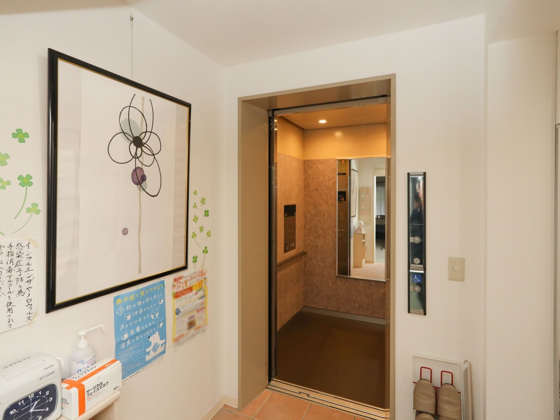 エレベーターに鏡や手すりがある。エレベーター前の壁には、絵画やお知らせの掲示物などが飾られている。