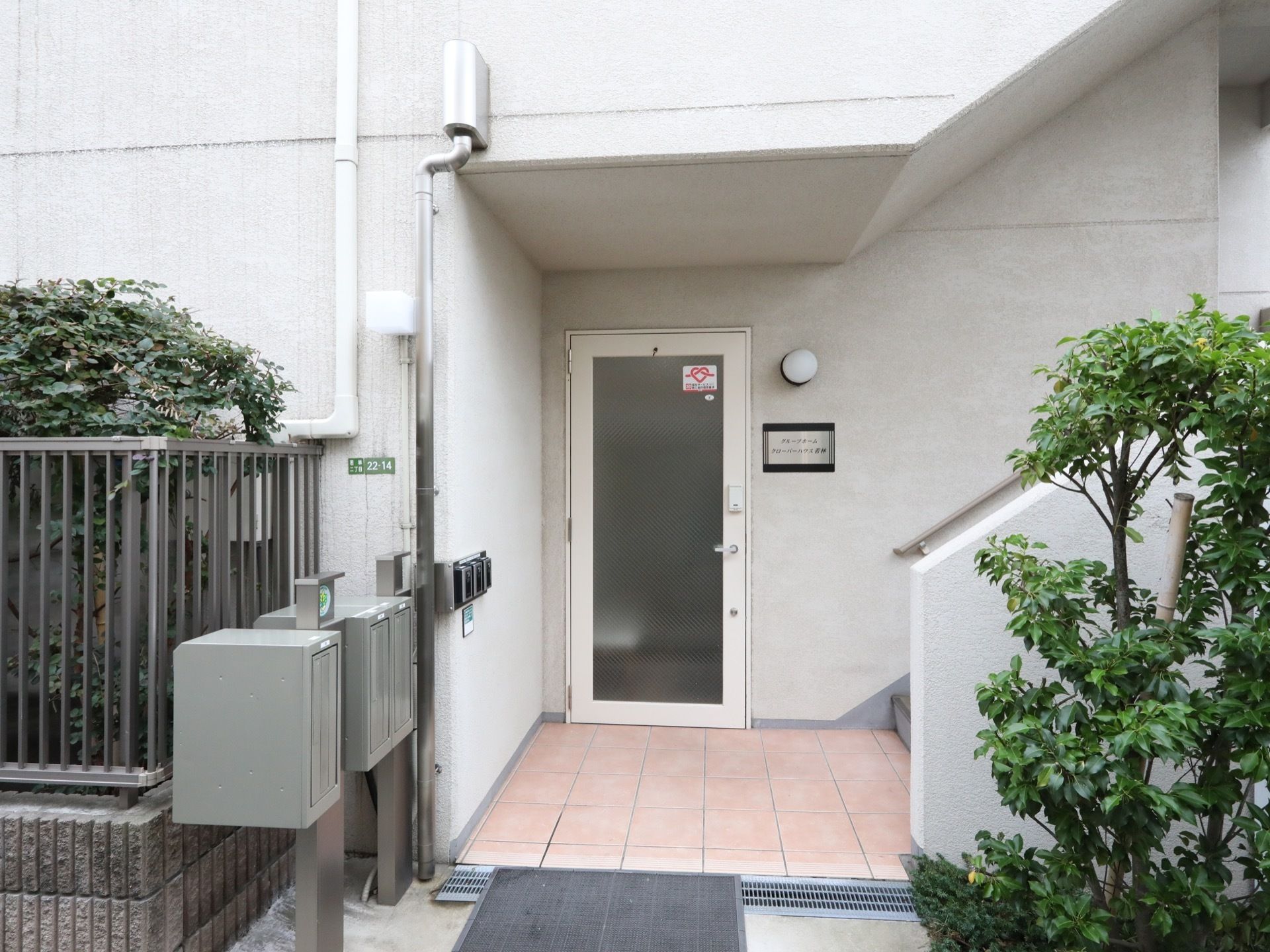 玄関のドアは白い枠組みにすりガラスがはめられている。ドアの横にライトと施設名がかかれた看板が設置されている。