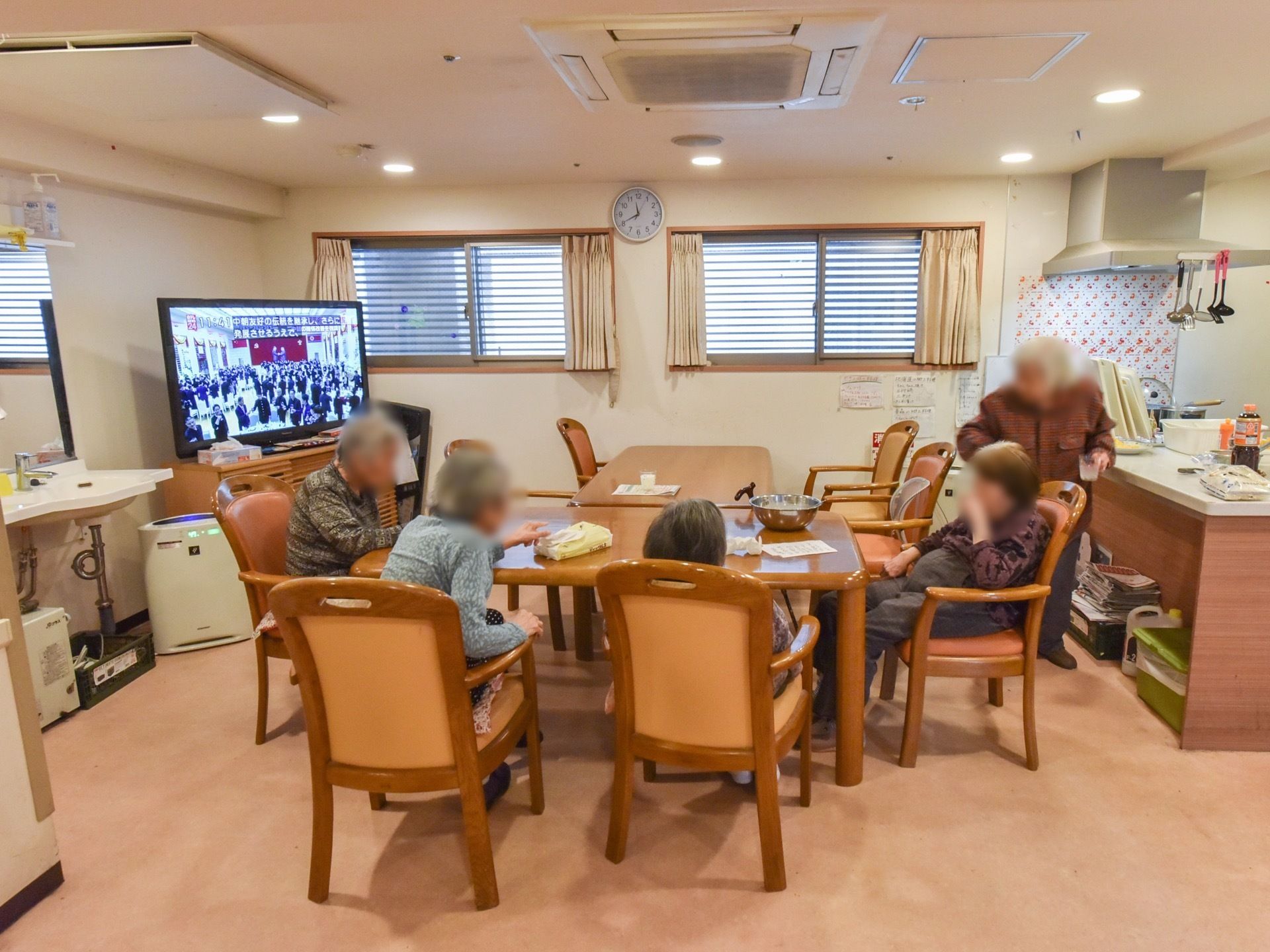 テーブルセットが配置され、テレビが映っている共有スペースでは、５名の女性の入居者様が椅子に腰掛け会話されている。