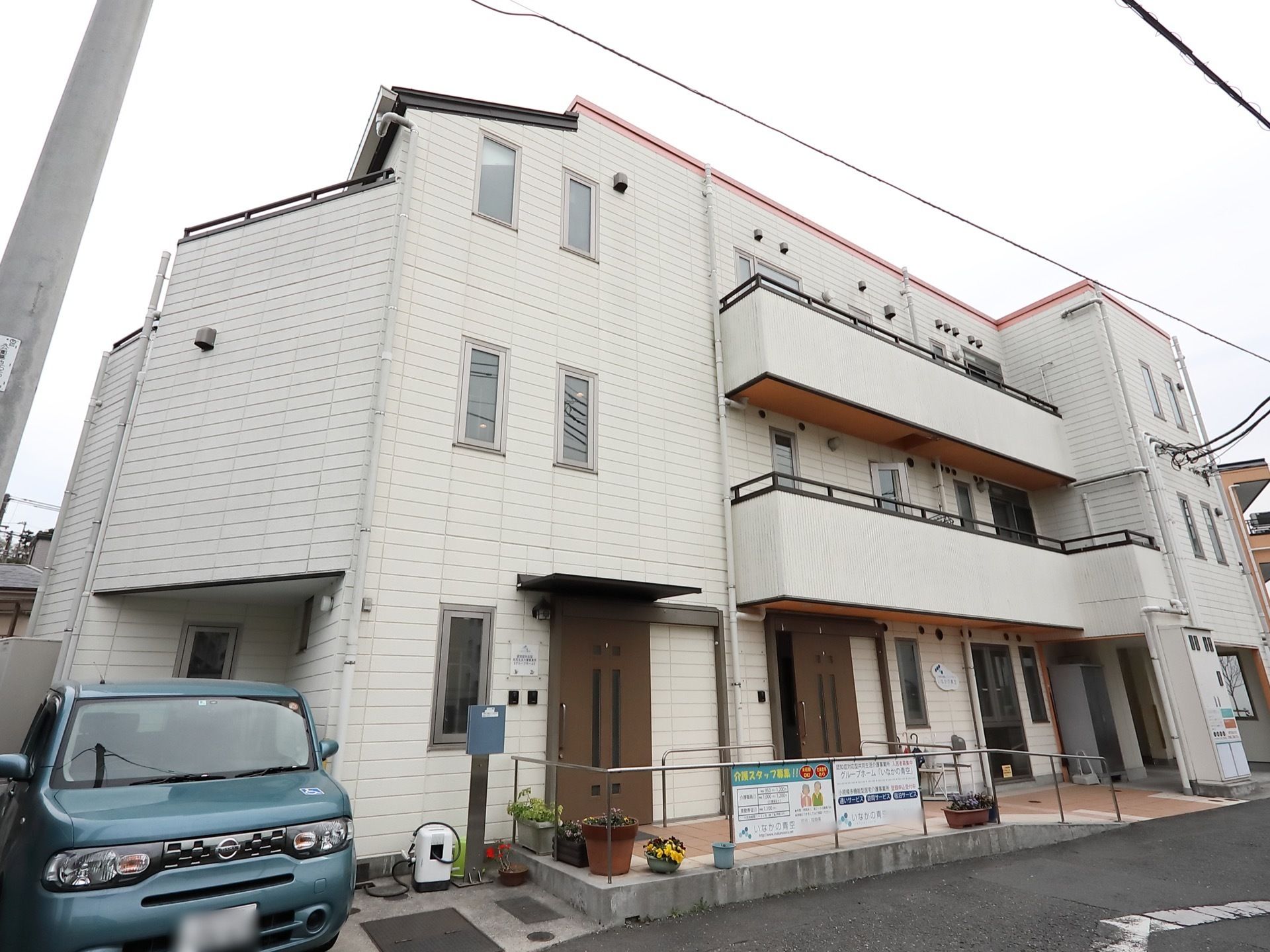 白い外壁の３階建ての建物である。階段部分の縦長の窓が並んでいる。施設の前には自動車が１台停まっている。